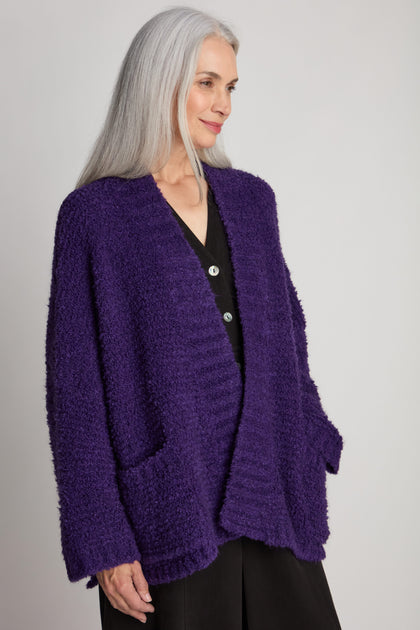 A person with long gray hair wears a Boucle Knit Cable Trim Cardi and stands against a plain background, looking to their left.