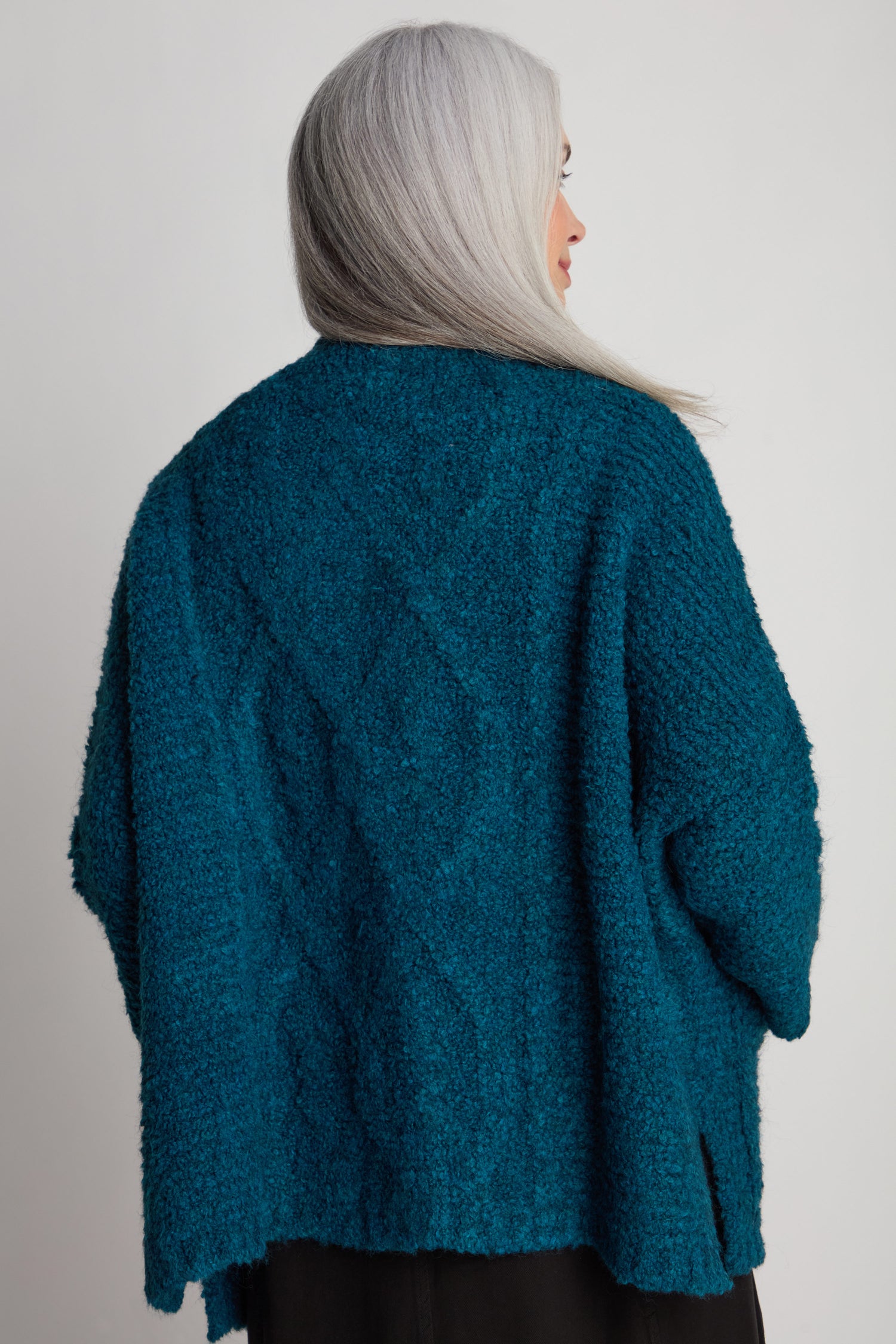 A person with long gray hair is seen from the back, wearing a teal Boucle Knit Cable Trim Cardi over a black outfit against a neutral background.