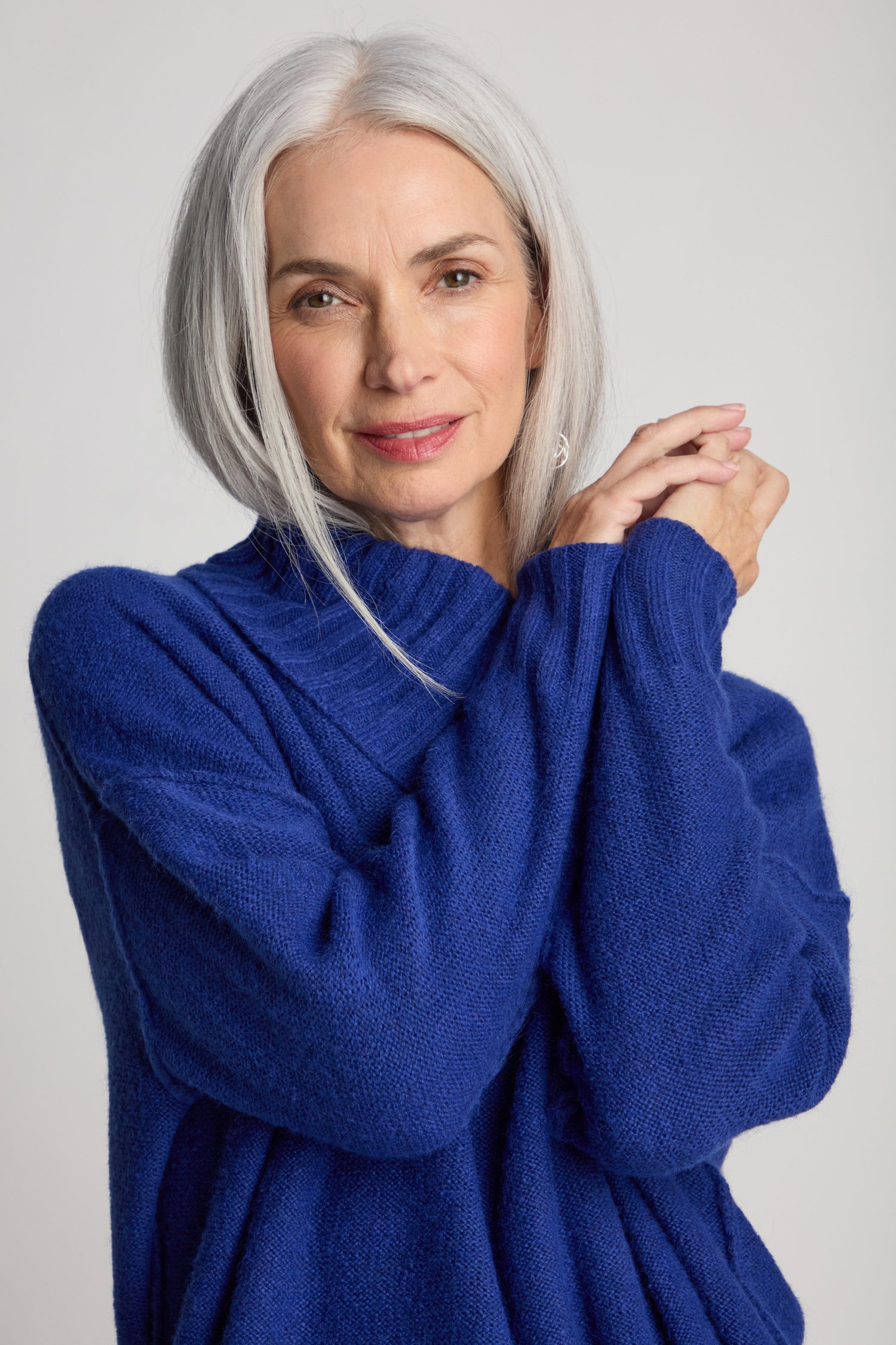 A woman with long gray hair wearing a blue Wool Viscose Mix Asymmetric Jumper stands with her hands clasped, looking at the camera.