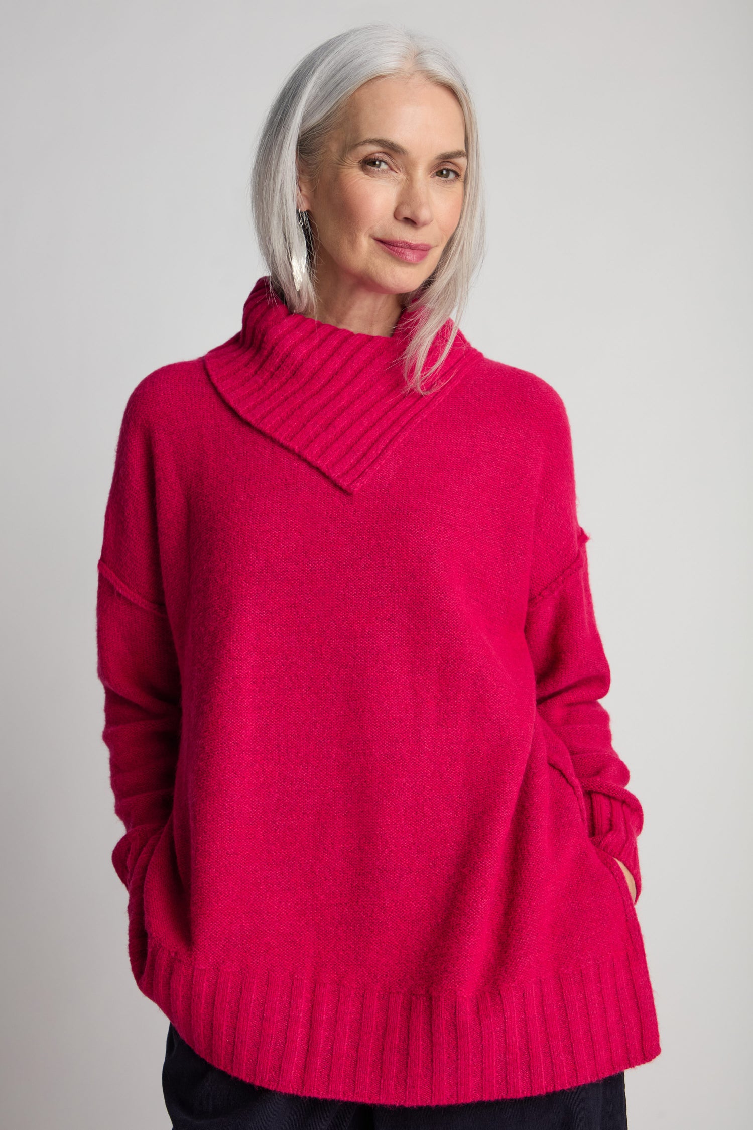 A woman with gray hair is wearing a bright pink Wool Viscose Mix Asymmetric Jumper with a folded collar, her hands tucked into the pockets. She stands against a plain background, embodying contemporary style.