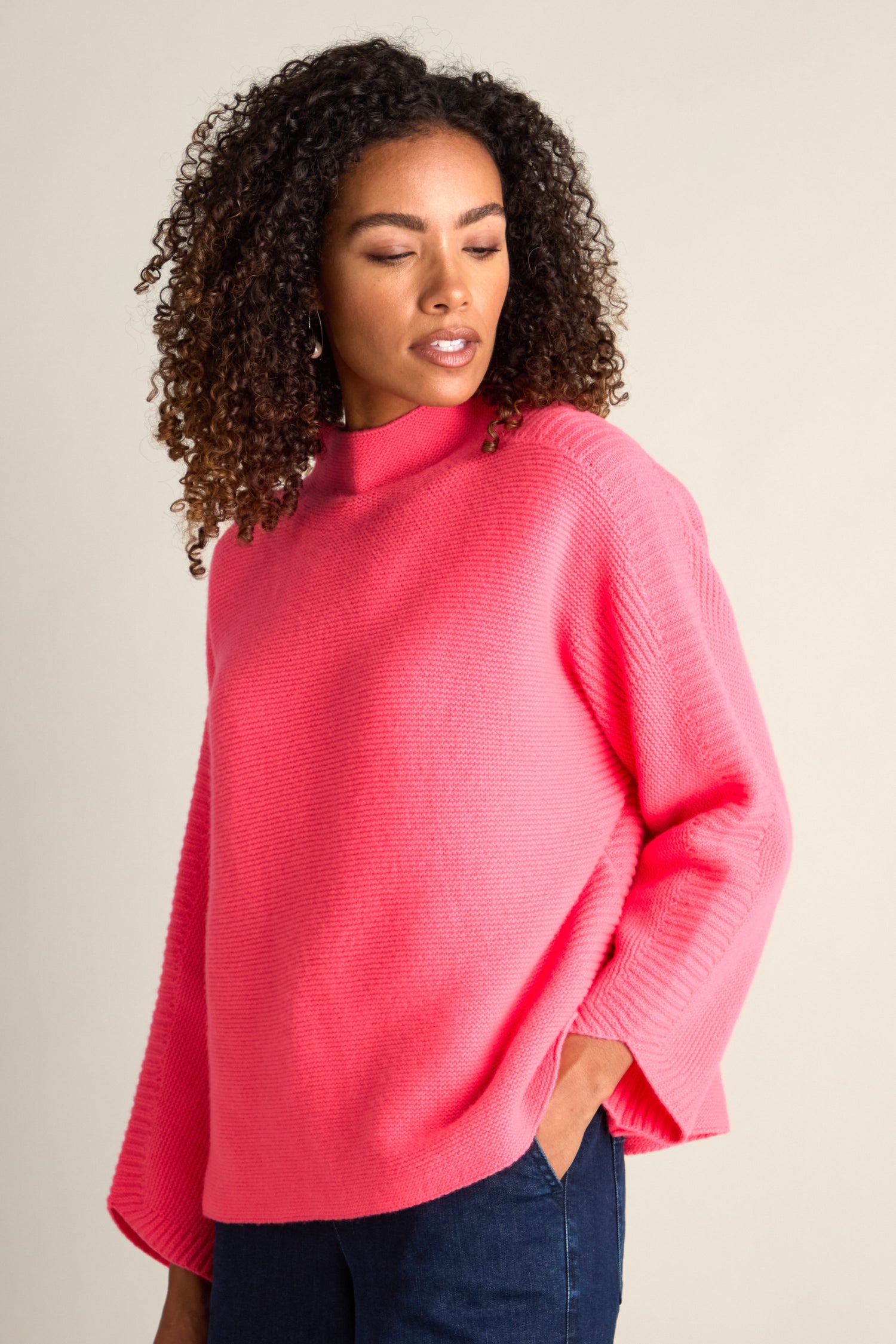 A person with curly hair wearing a cozy Merino Wool Stitch Detail Jumper and blue jeans stands against a plain background.