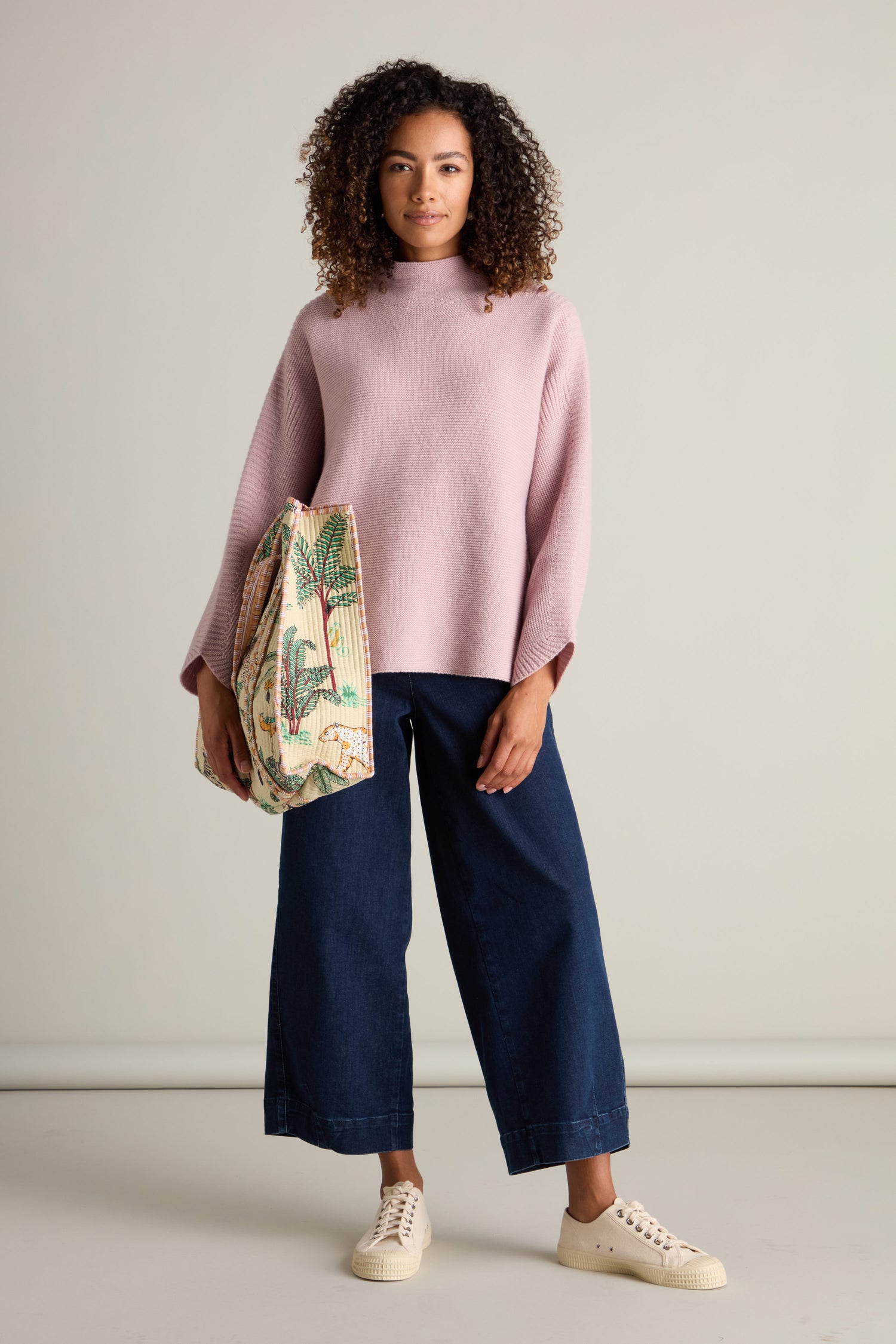 A person stands against a plain background wearing a cozy pink Merino Wool Stitch Detail Jumper, blue wide-leg jeans, and white sneakers, holding a patterned tote bag.