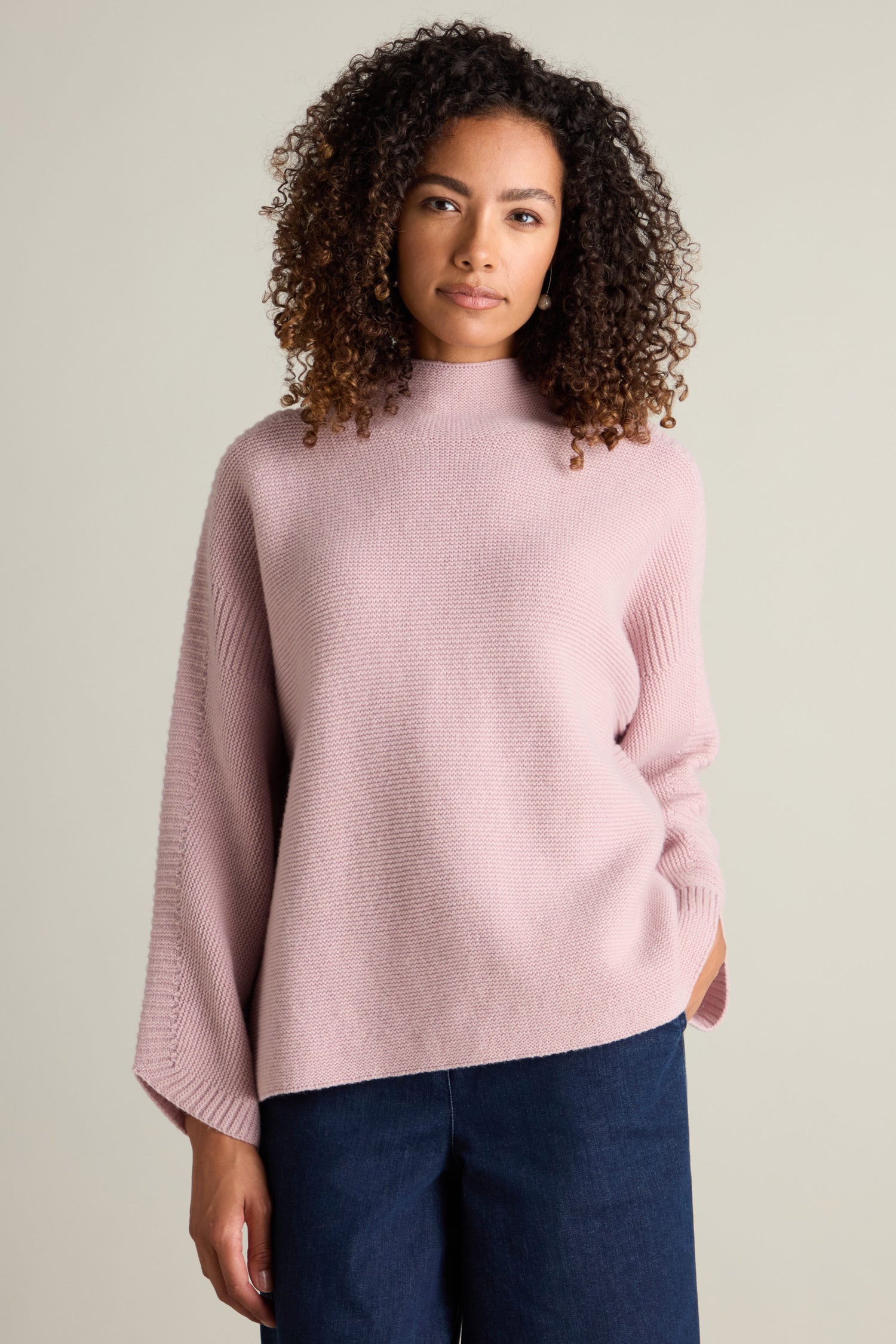 A person with curly hair wears a cozy Merino Wool Stitch Detail Jumper in pink and blue pants, standing against a plain background.