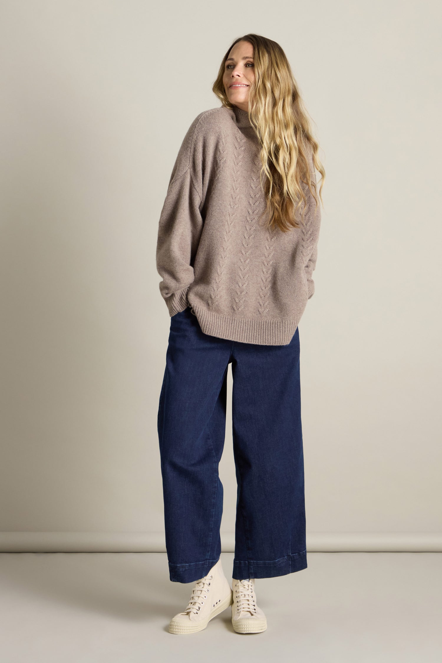 A person stands against a plain background, wearing a loose, light brown Cashmere Blend Cable Jumper, wide-leg blue pants, and white high-top sneakers. They have long, wavy blonde hair and are smiling slightly.