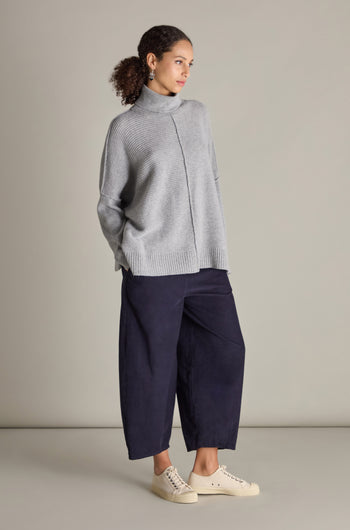 Against a plain background, a person stands wearing a grey Merino Wool Boxy Jumper, navy blue wide-leg pants, and white sneakers. Their curly hair is pulled back and they are looking to the side.