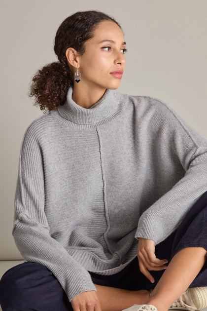 A person with curly hair tied back, wearing a grey Merino Wool Boxy Jumper and dark pants, sits against a plain background, looking to the side. They are also wearing earrings.