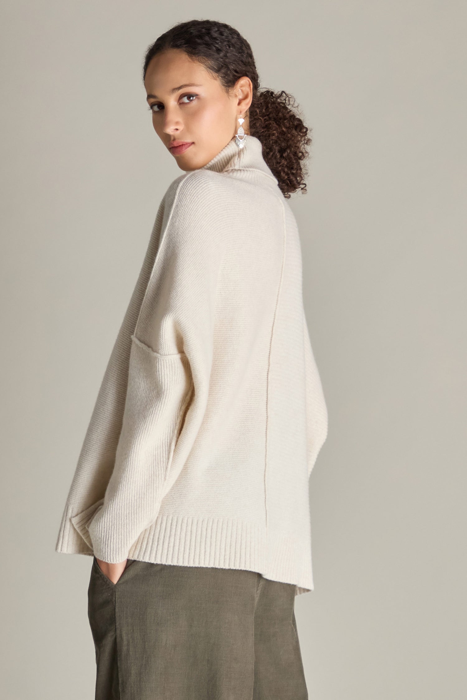 A woman with curly hair, wearing a cream-colored Merino Wool Boxy Jumper and olive pants, poses sideways against a plain background.