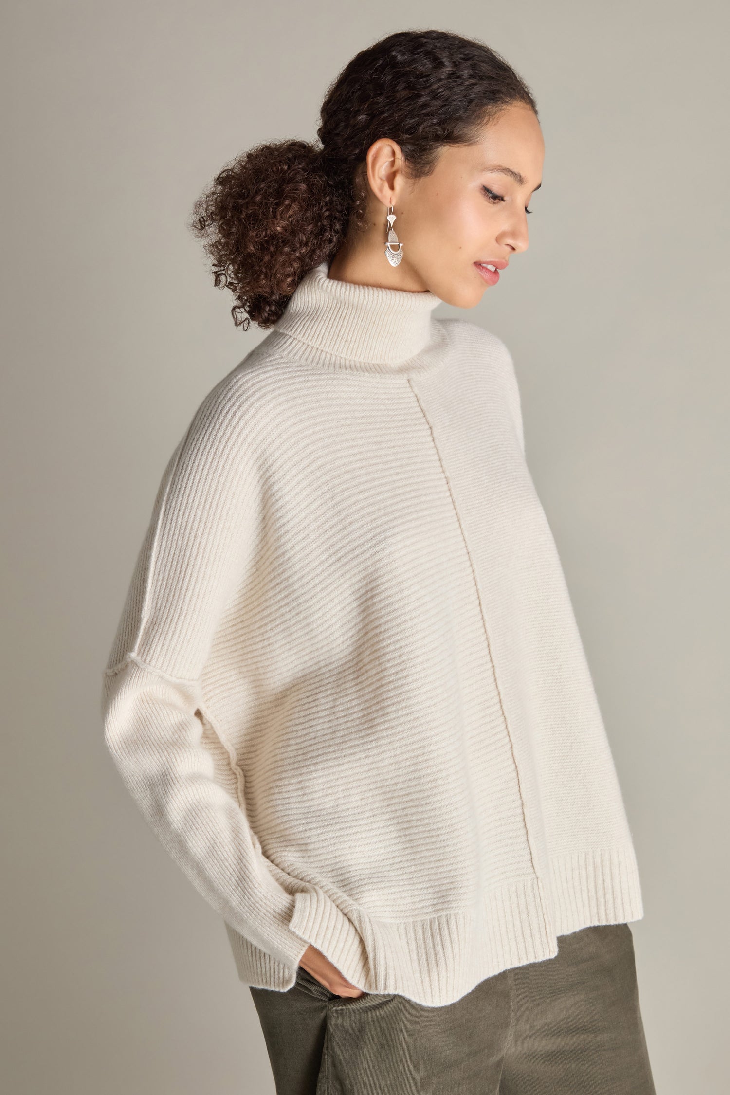 A person with curly hair wearing a light-colored Merino Wool Boxy Jumper and dangling earrings stands against a plain background with hands in pockets, looking to the side.