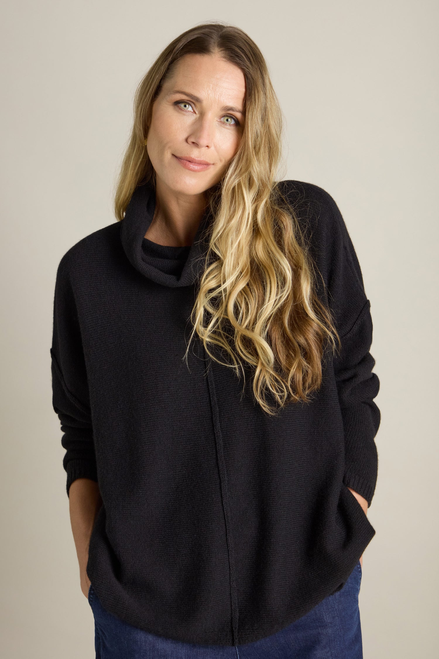 A person with long wavy hair, wearing a dark Merino Wool Cowl Neck Jumper, stands against a plain background with their hands in their pockets.