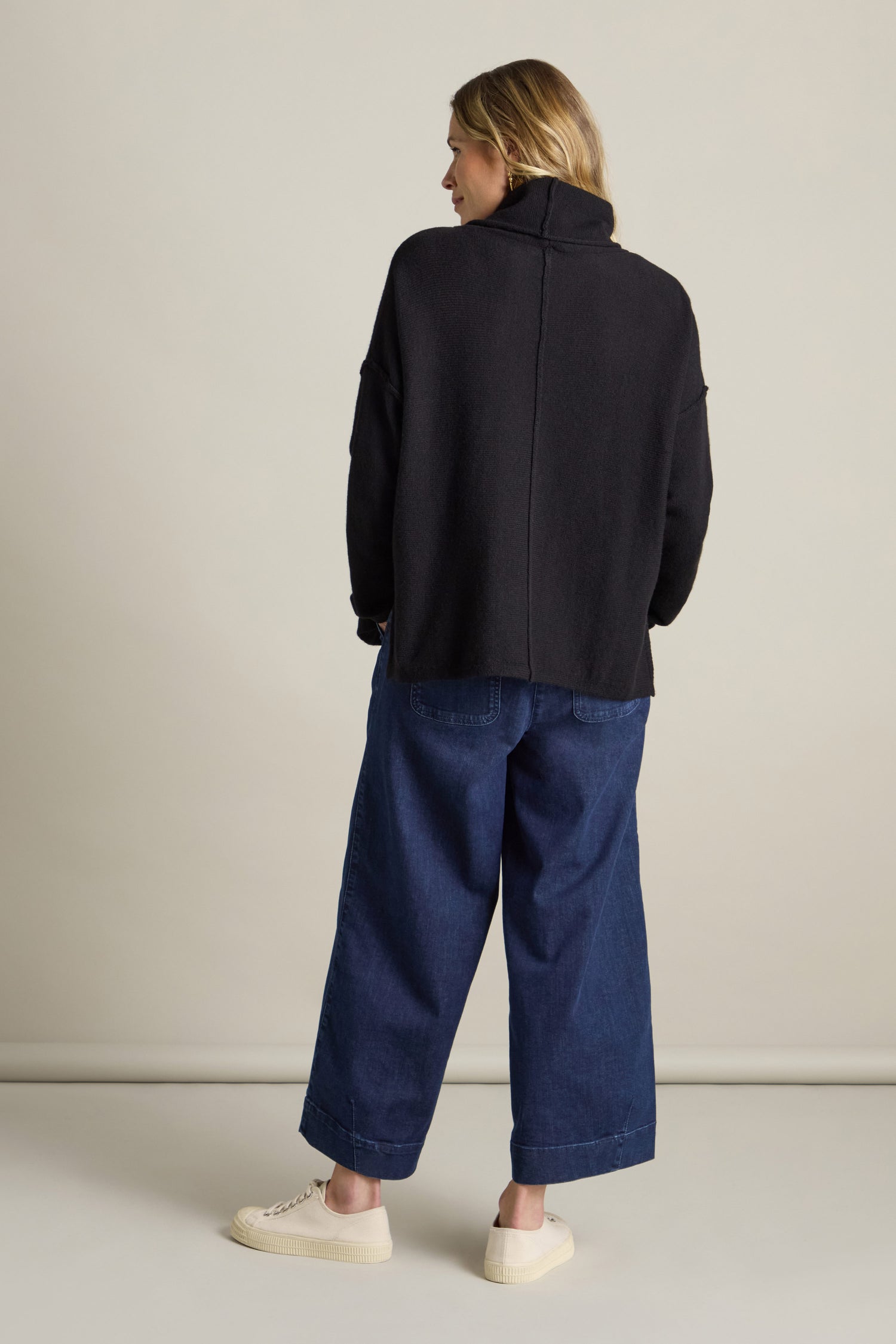 Person with shoulder-length hair wearing a black Merino Wool Cowl Neck Jumper, blue wide-leg jeans, and white shoes, standing and facing away in front of a neutral background.