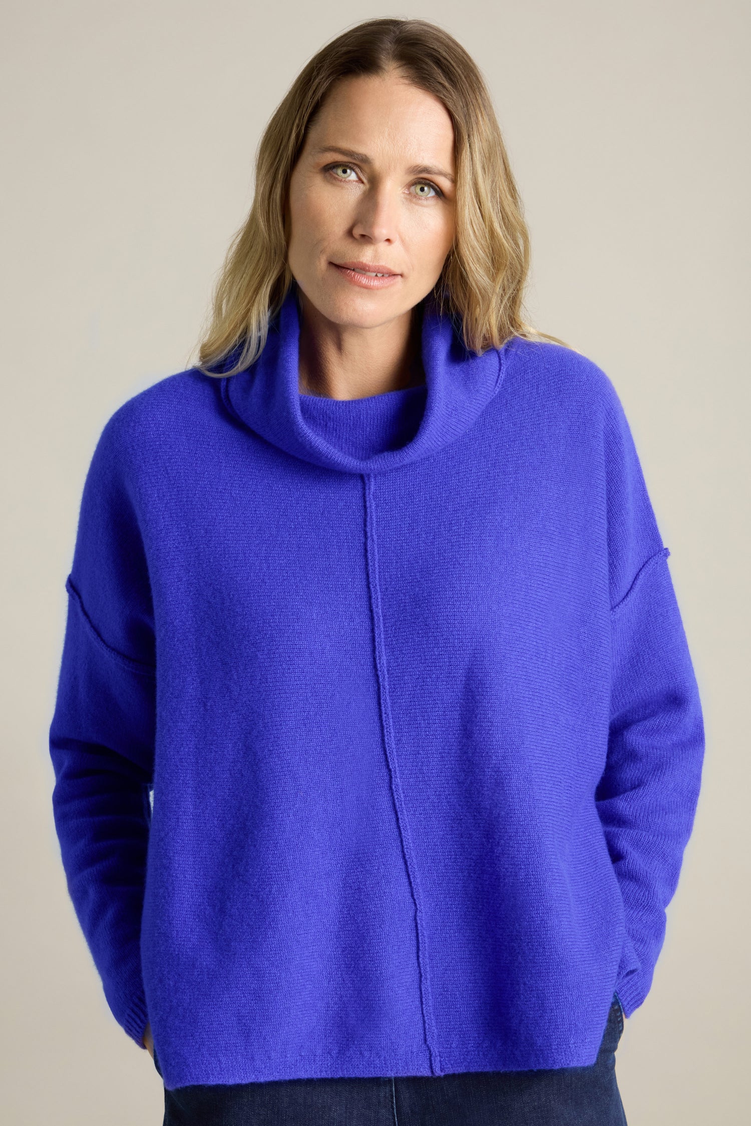 A person with long hair wearing a blue Merino Wool Cowl Neck Jumper stands against a neutral background, looking into the camera.