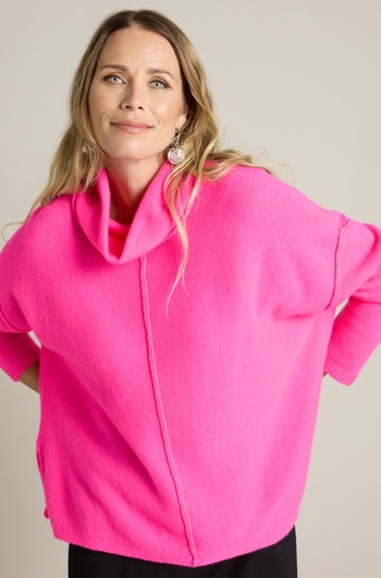 A person stands in a relaxed pose, wearing a bright pink Merino Wool Cowl Neck Jumper and black pants against a plain background.