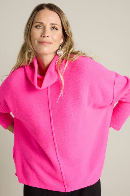 A person stands in a relaxed pose, wearing a bright pink Merino Wool Cowl Neck Jumper and black pants against a plain background.