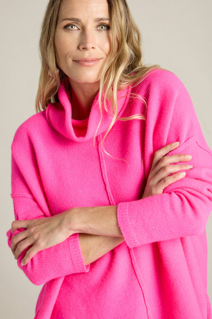 A person with long hair wearing a bright pink Merino Wool Cowl Neck Jumper stands with arms crossed, smiling slightly at the camera.