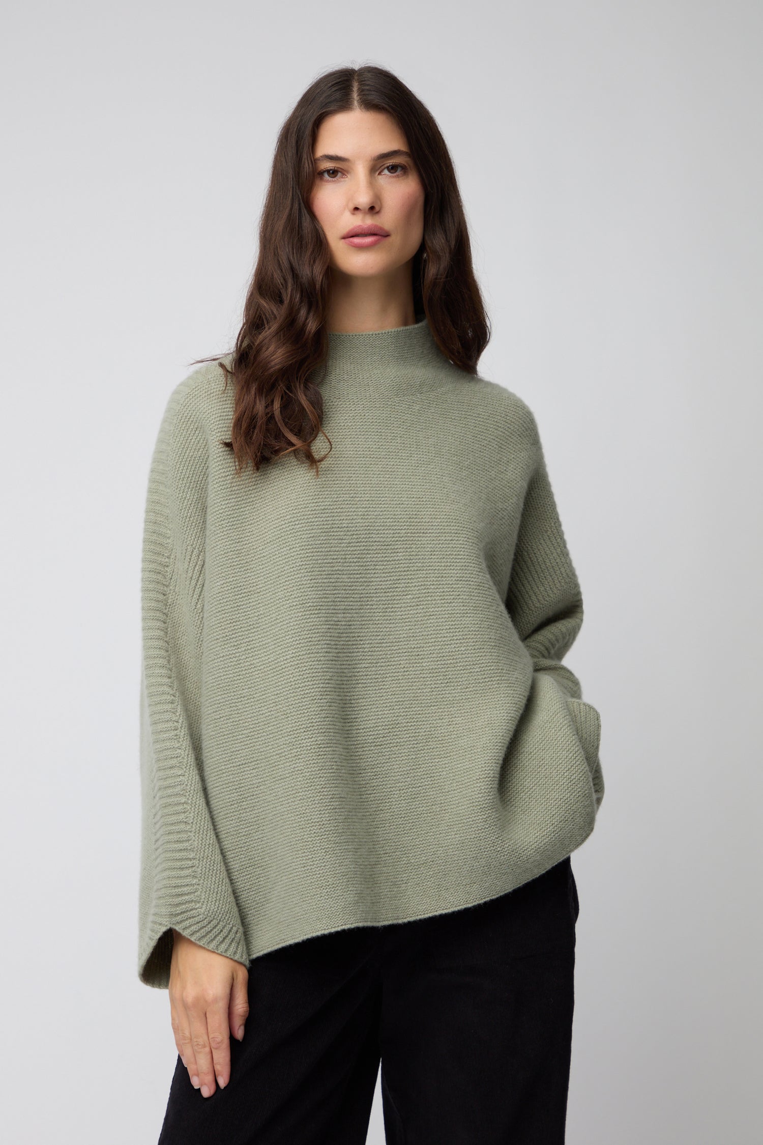 A woman with long brown hair wears the Merino Wool Stitch Detail Jumper, a loose, light green knit sweater, paired with black pants against a plain gray background.