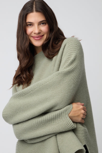 A woman with long brown hair smiles at the camera, wearing the Merino Wool Stitch Detail Jumper in light green—an oversized knitwear staple—set against a plain background.