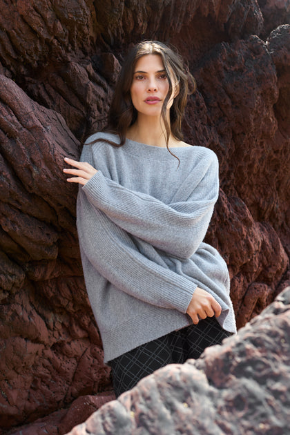 A woman with long dark hair wears the Merino Rib Detail Jumper and plaid pants, standing against a backdrop of dark, textured rocks.