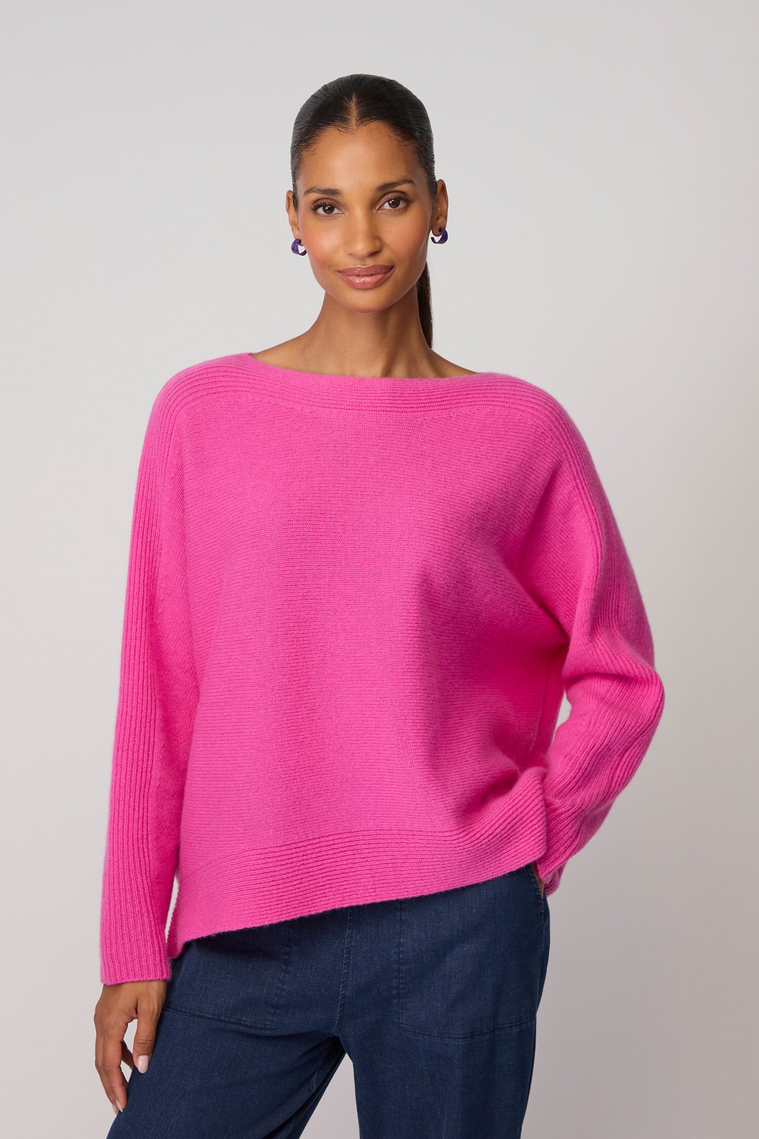 A woman stands against a plain background, wearing the Merino Rib Detail Jumper in bright pink and dark blue pants, with her hand in her pocket.