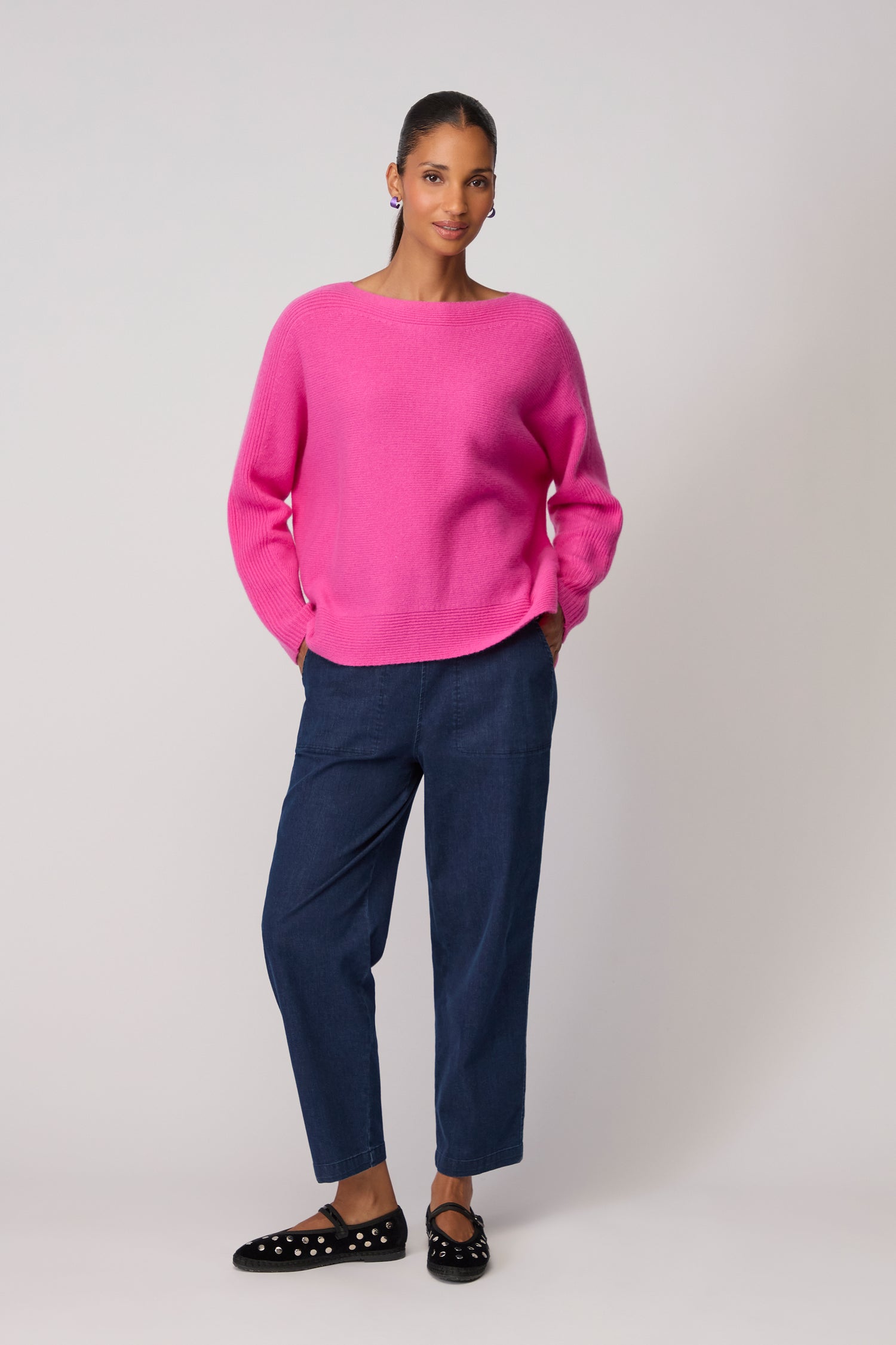 A woman stands against a light background wearing a bright pink Merino Rib Detail Jumper, blue relaxed-fit pants, and black flat shoes with white spots.