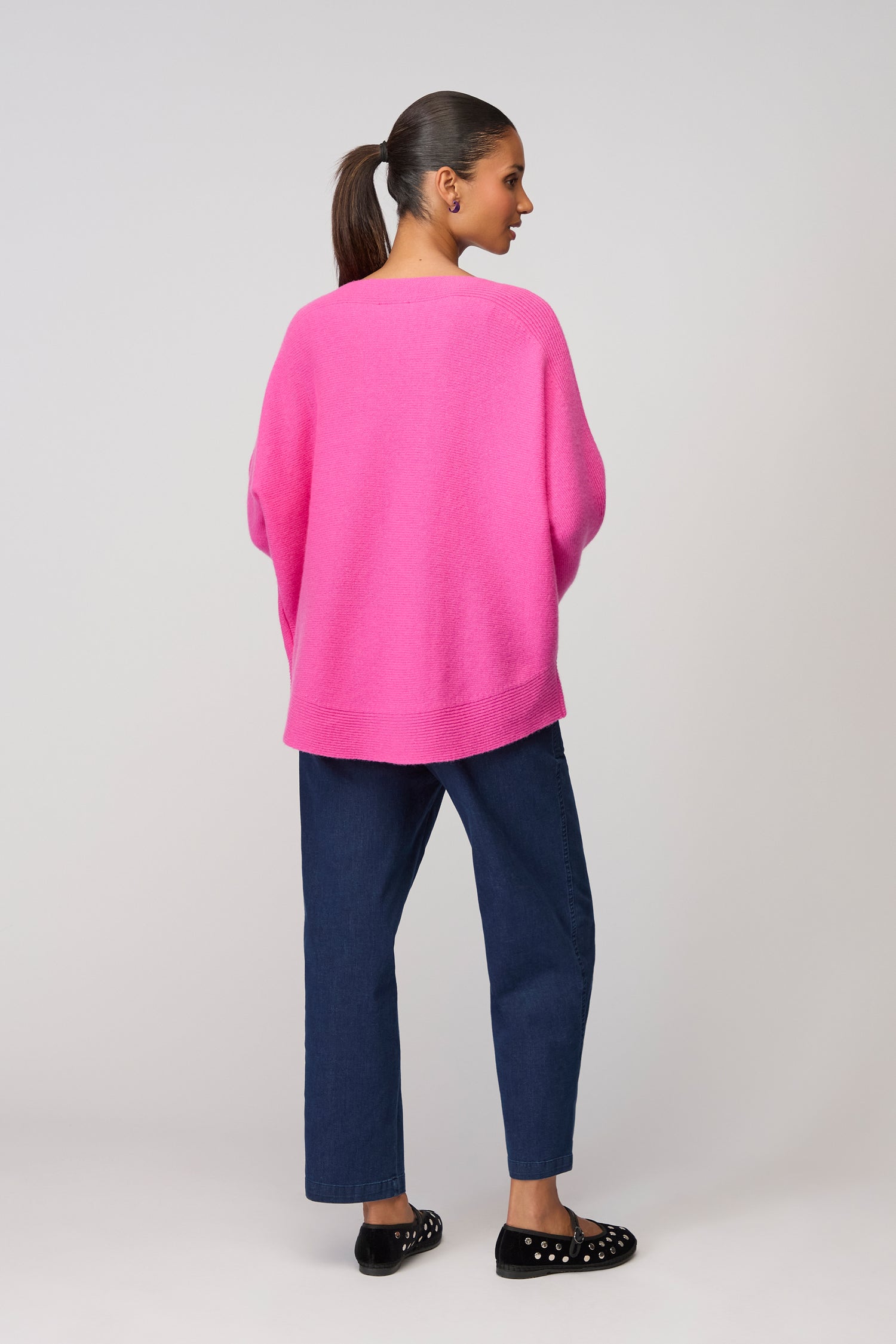 A woman stands facing away, wearing a bright pink Merino Rib Detail Jumper, dark blue jeans, and black polka dot shoes against a plain background.