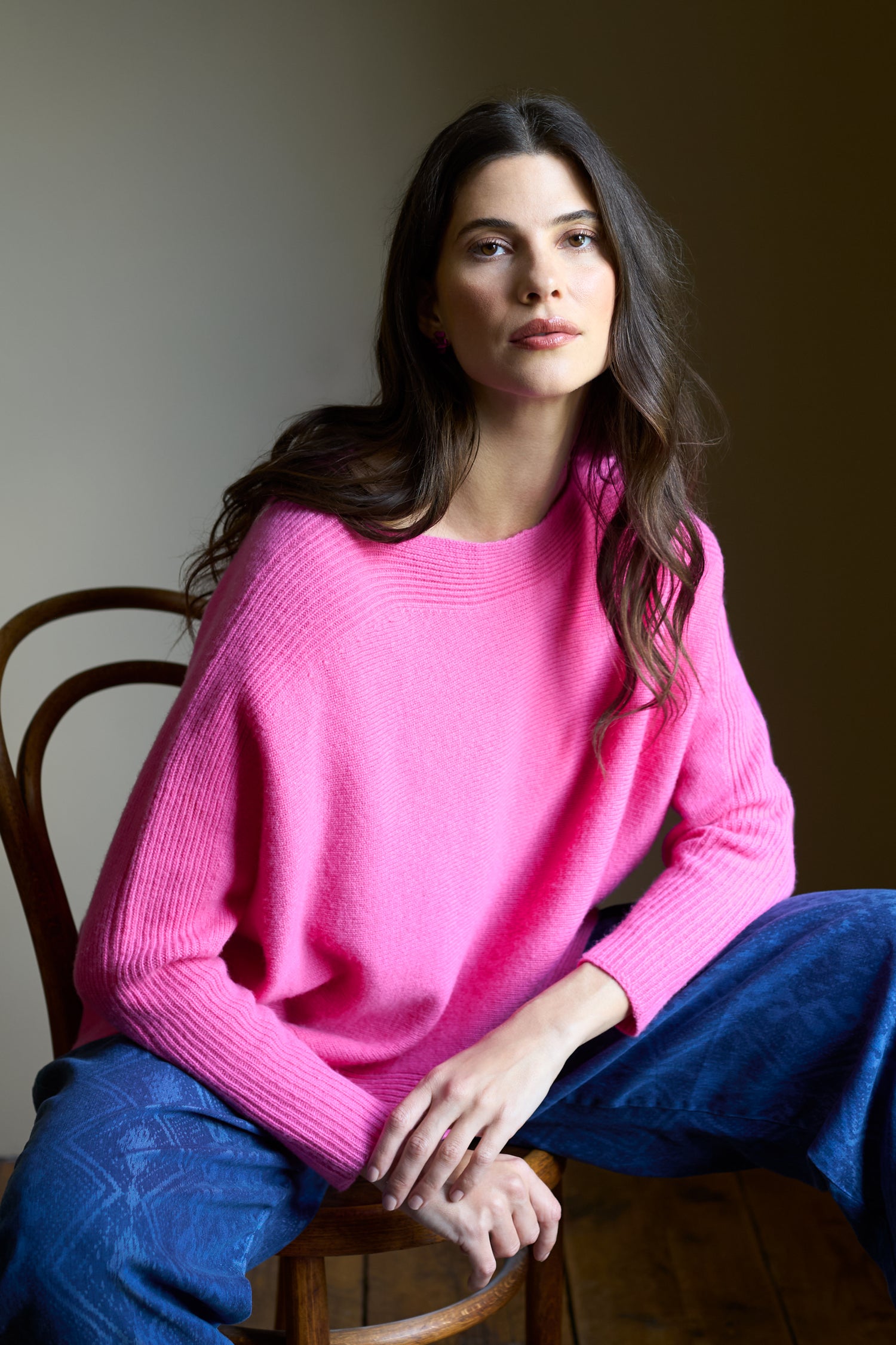 A woman with long brown hair sits on a wooden chair against a plain background, wearing the bright pink Merino Rib Detail Jumper and blue patterned pants.