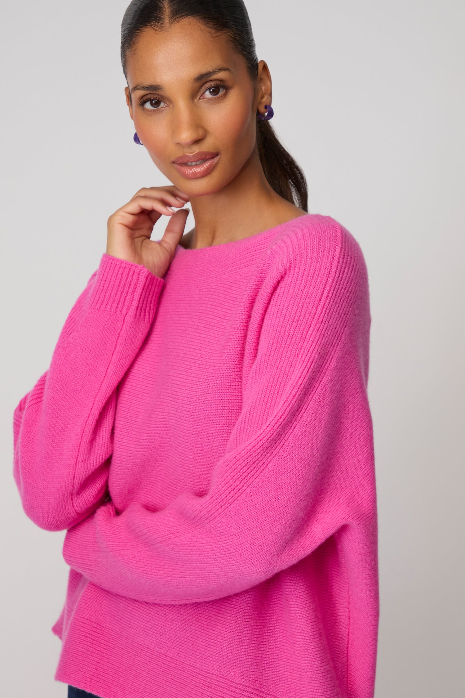 A woman wearing a bright pink Merino Rib Detail Jumper and blue earrings stands against a plain background, looking at the camera with her hand near her face.