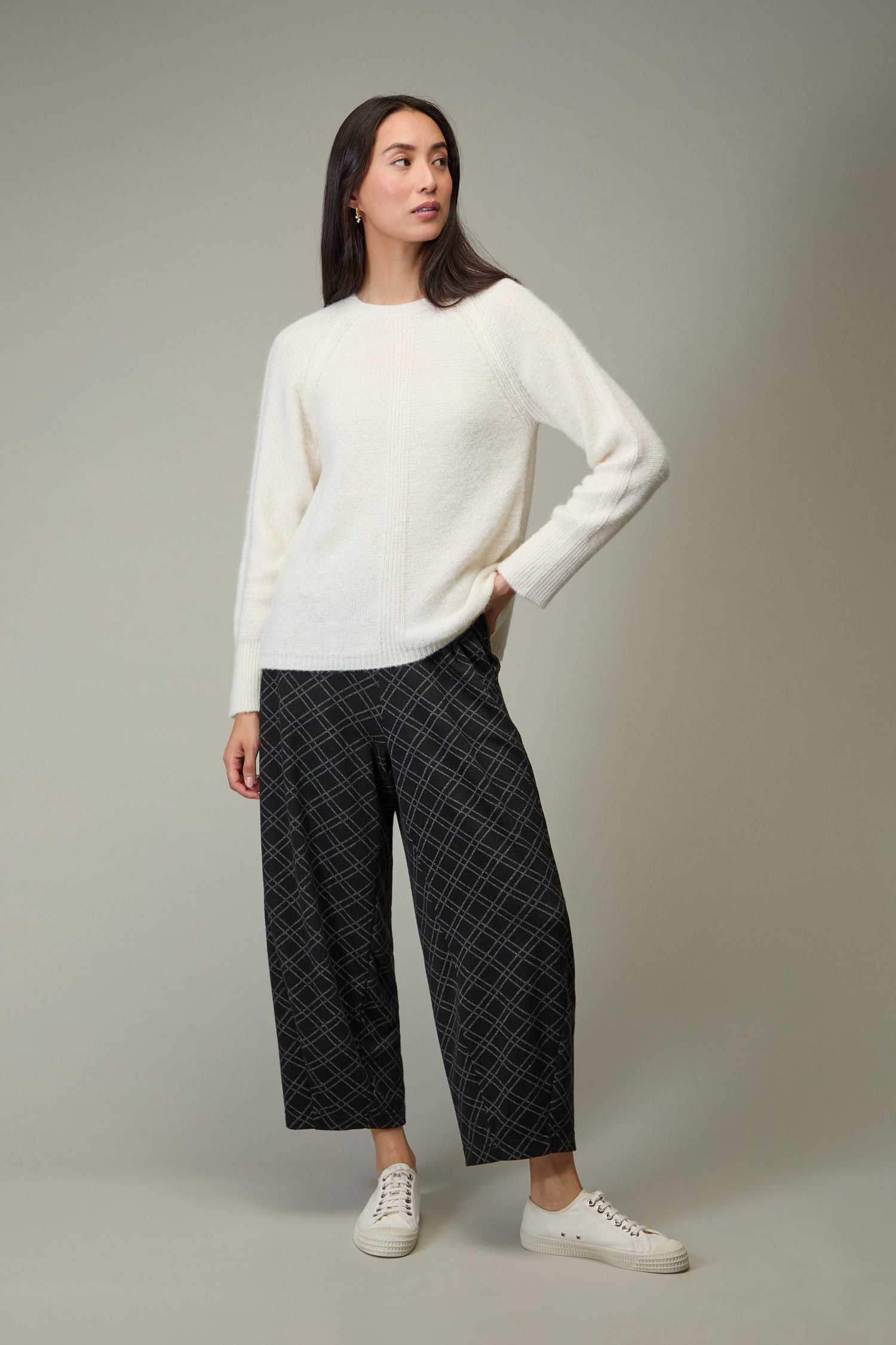 A woman stands against a plain background wearing the Alpaca Blend Seam Detail Jumper, black patterned wide-leg pants, and white sneakers—an effortlessly modern take on quiet luxury.