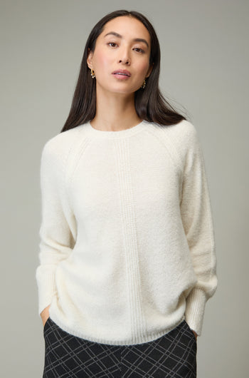A woman stands against a plain background with her hands in her pockets, wearing an Alpaca Blend Seam Detail Jumper and patterned dark pants.