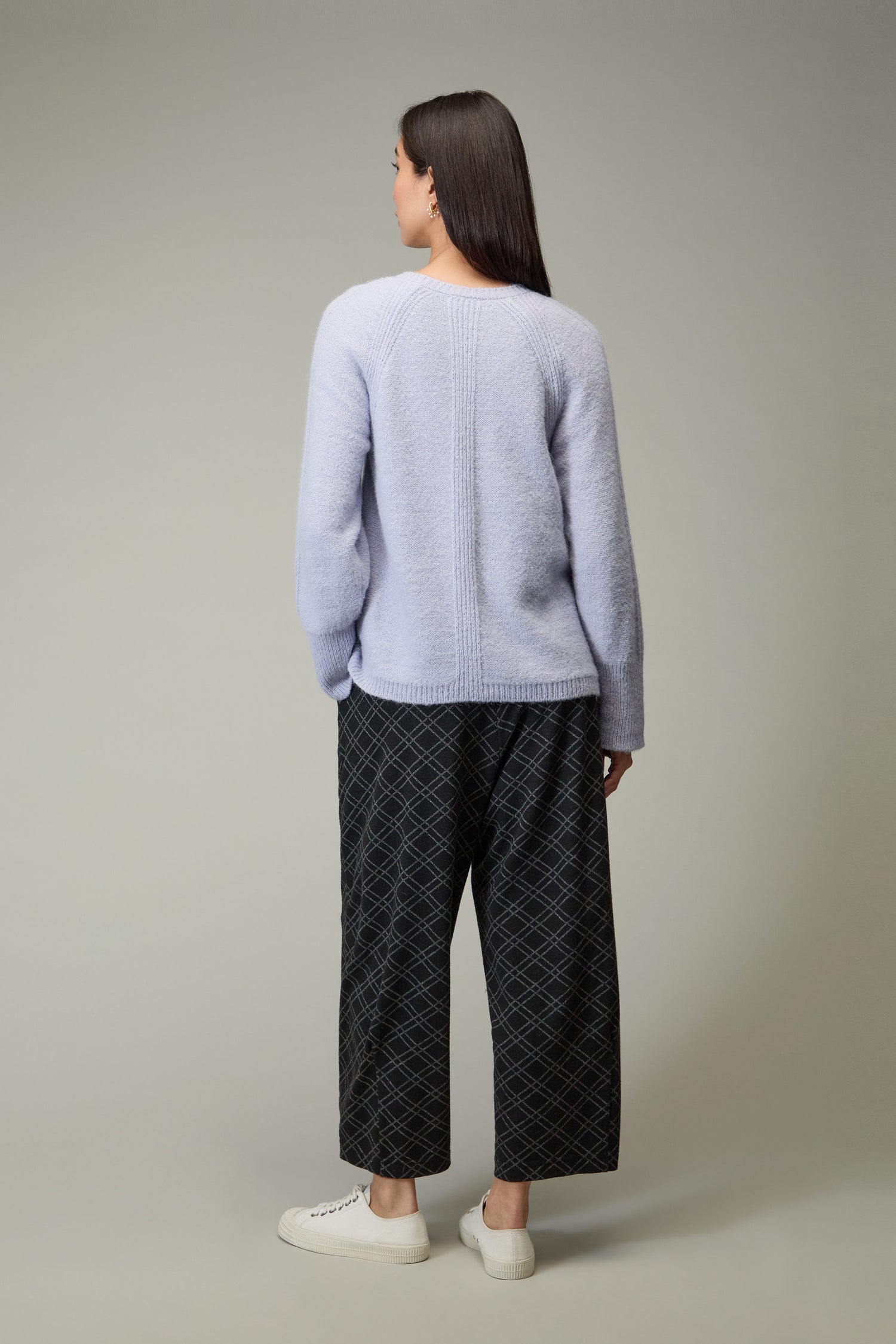 Woman stands with her back to the camera, wearing the Alpaca Blend Seam Detail Jumper in light purple, dark patterned wide-leg pants, and white sneakers against a plain backdrop—a perfect nod to quiet luxury.
