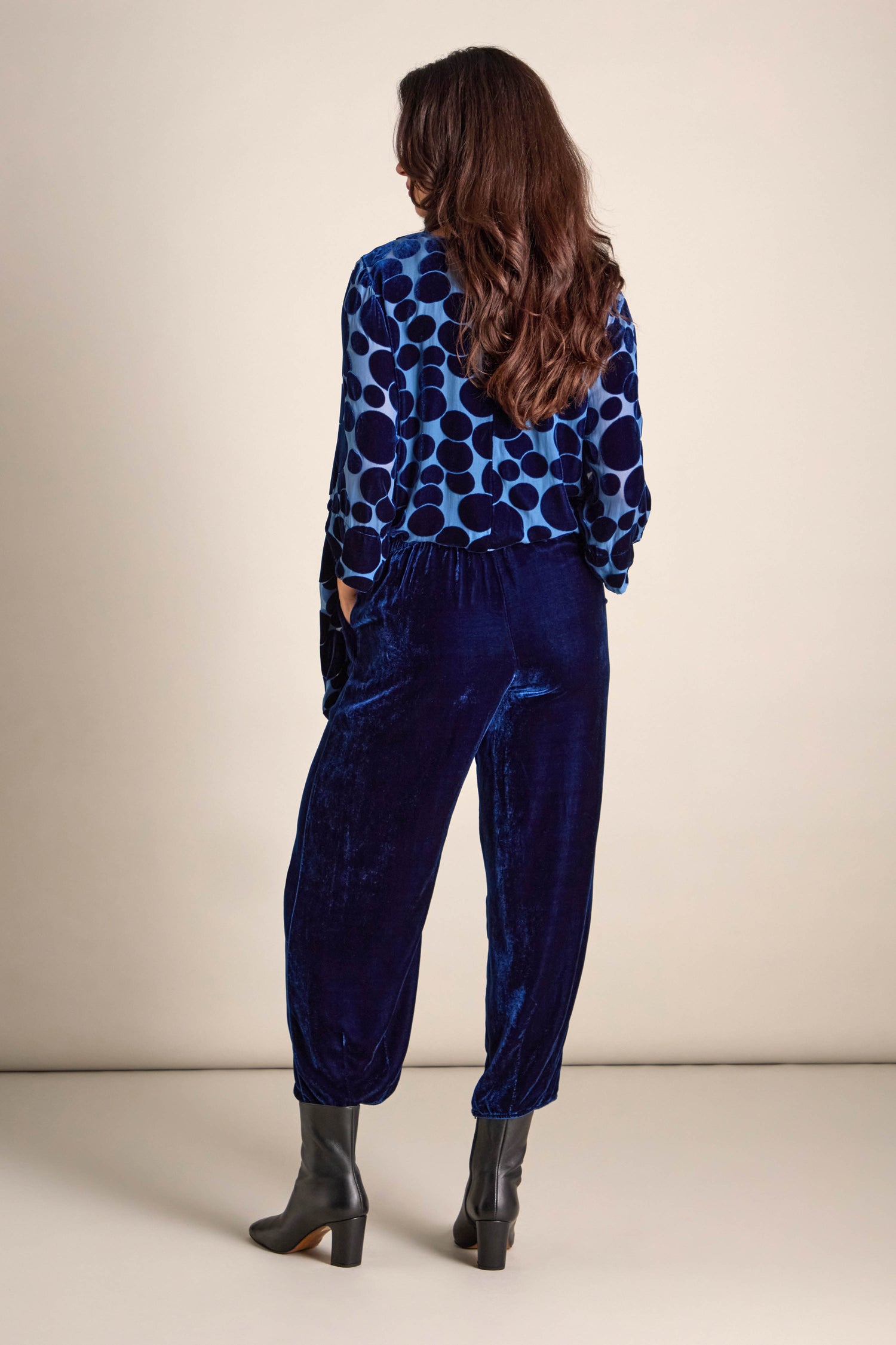 A woman wearing a blue polka dot top and dark blue Silk Velvet Trousers with an elasticated waistband stands facing away. Her long, wavy hair flows down, pairing perfectly with her chic black ankle boots.