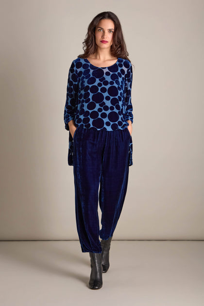 A woman wearing a blue polka dot top and the Silk Velvet Trousers, featuring an elasticated waistband, stands with her hands in the pockets against a neutral background.