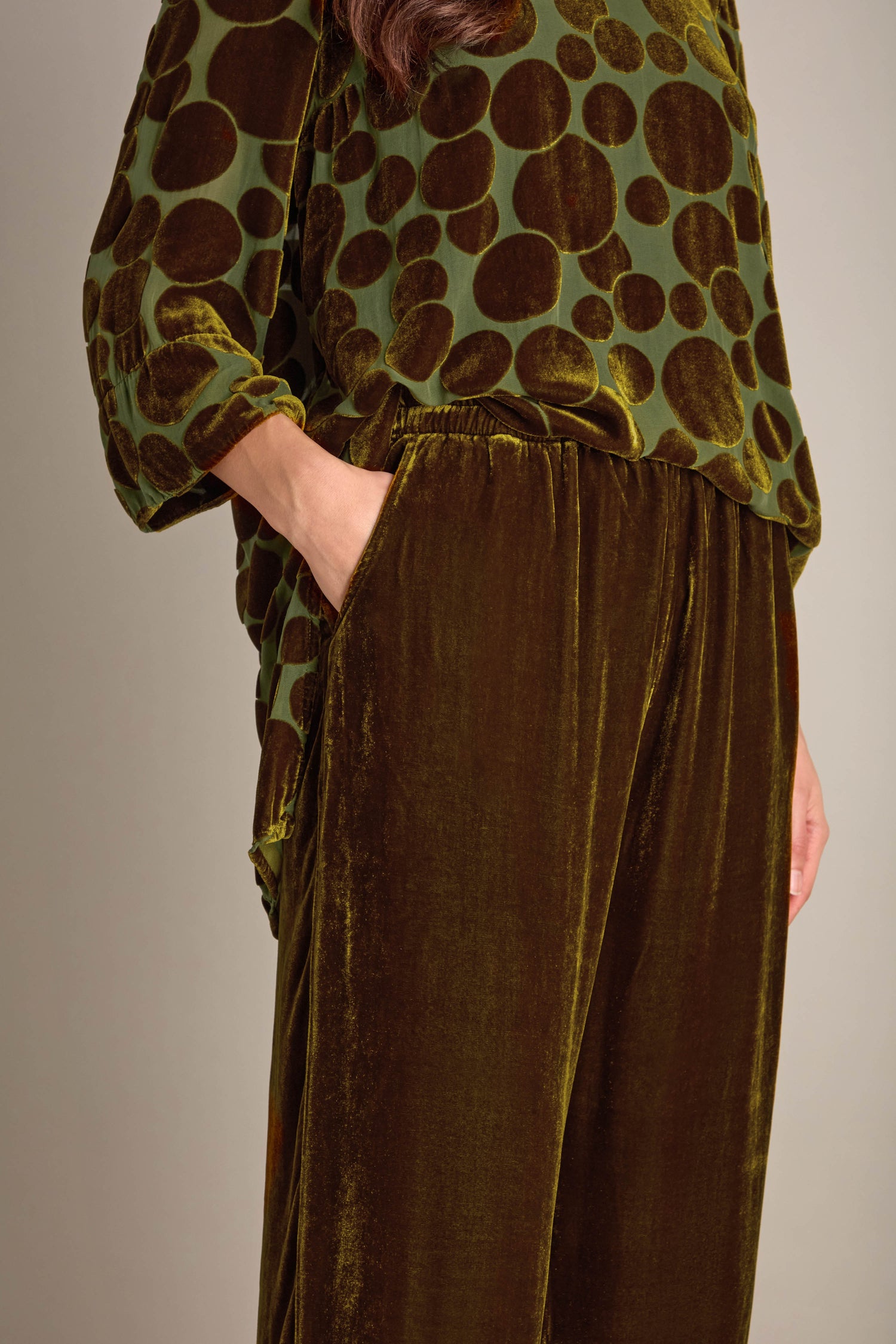 A person wearing a green ensemble with dark circular patterns on the top, paired with Silk Velvet Trousers that have an elasticated waistband. Their left hand is in one of the trousers' pockets.