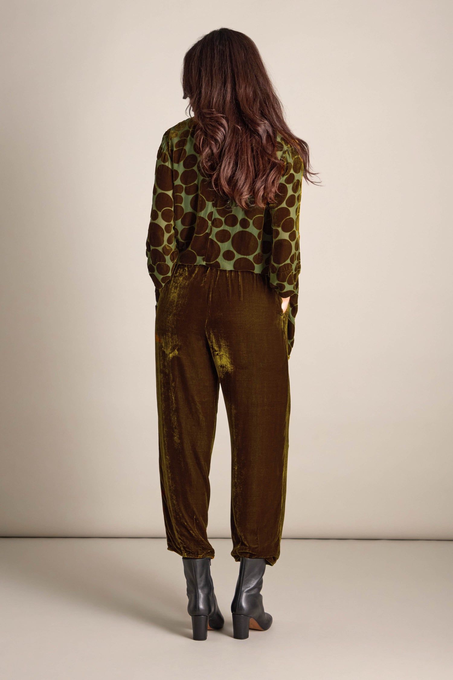 A woman with long brown hair stands with her back to the camera, dressed in a green polka dot blouse, Silk Velvet Trousers that feature an elasticated waistband, and black heeled boots.