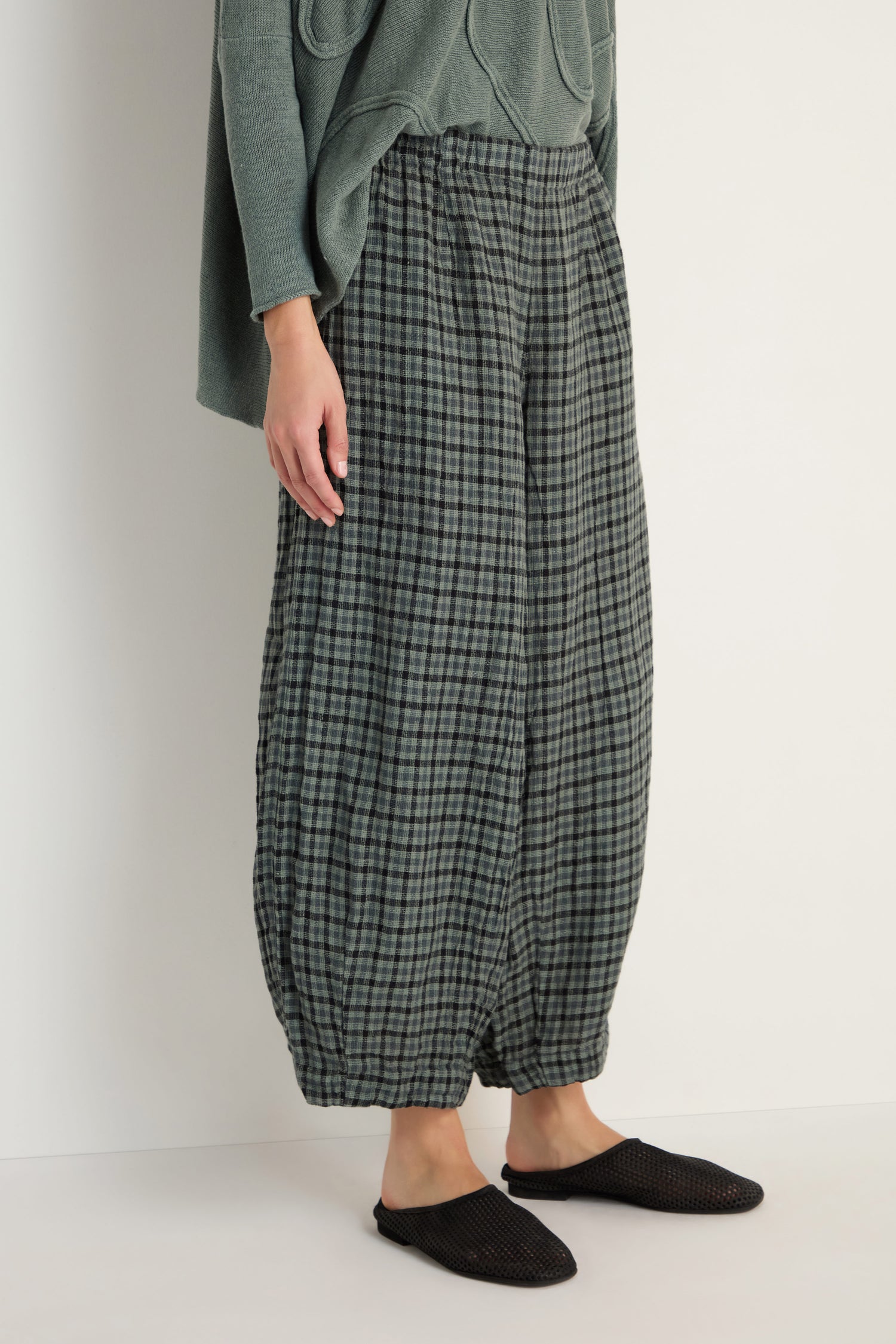 Wearing Check Linen Bubble Trousers in green and grey with a loose silhouette, paired with an olive green top and black slip-on shoes, standing against a plain light background.