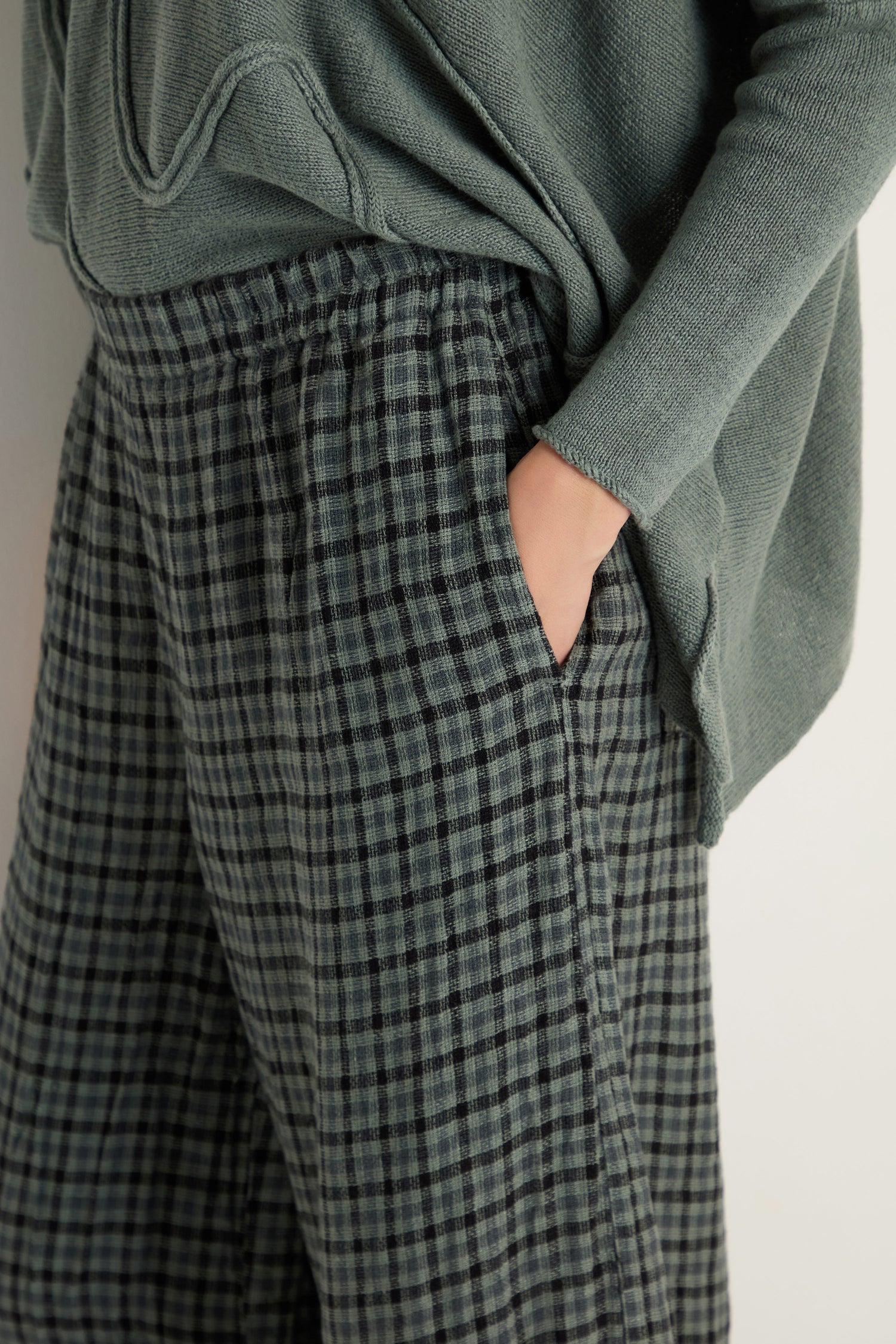 Model wears Check Linen Bubble Trousers and a sage green sweater, showing the trousers' relaxed silhouette with one hand in pocket, photographed from the waist down.
