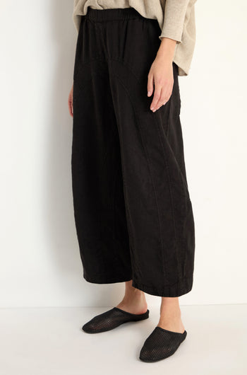 Model wears Curved Seam Linen Trousers in black with a relaxed leg, paired with a beige top and black slip-on shoes, against a plain white background.