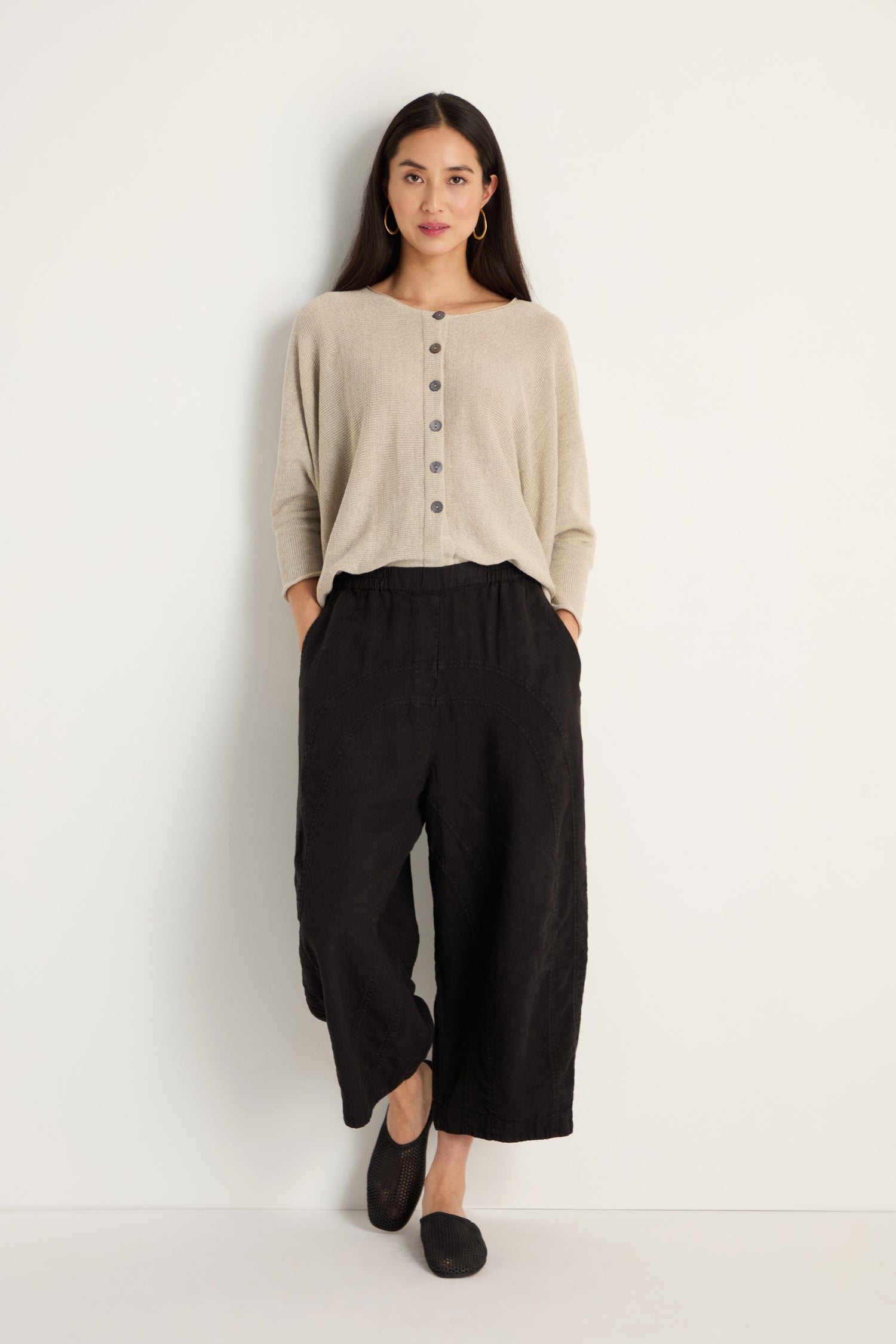 A woman stands against a white wall, wearing a beige buttoned top, black shoes, and Curved Seam Linen Trousers featuring relaxed legs and curved seam details.