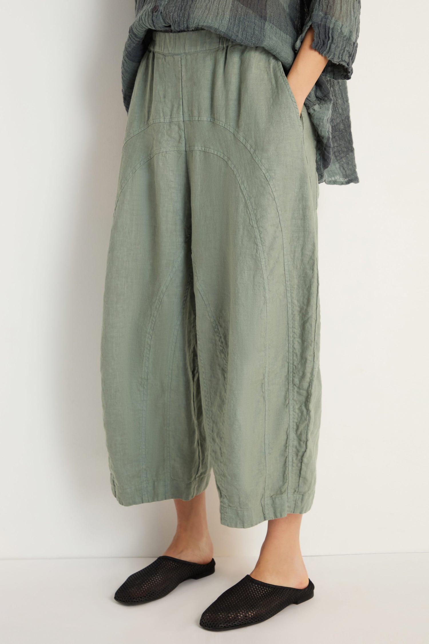 Lower body shot of a person wearing Curved Seam Linen Trousers in light green with relaxed legs, hands in pockets, paired with black slip-on shoes and a textured dark top.
