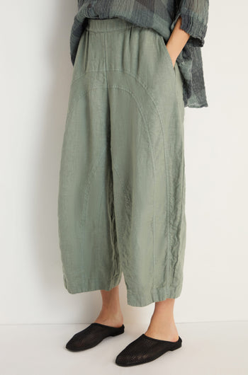 Lower body shot of a person wearing Curved Seam Linen Trousers in light green with relaxed legs, hands in pockets, paired with black slip-on shoes and a textured dark top.