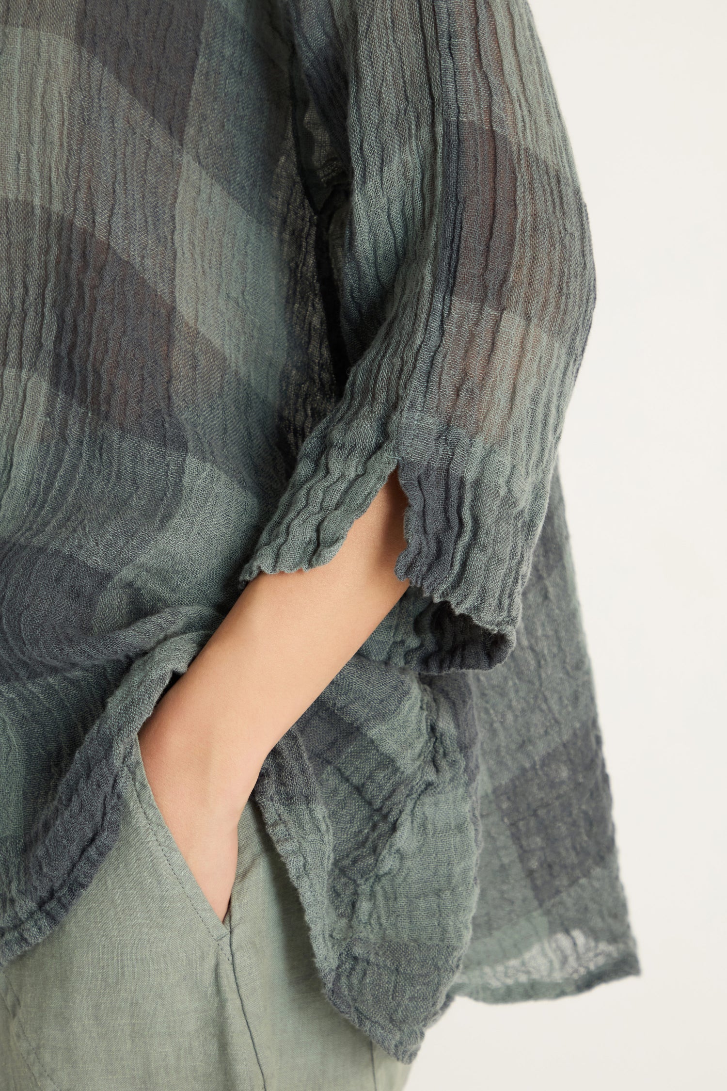 Wearing the Large Check Sage Linen Swing Shirt—a breathable, lightweight top with a relaxed swing silhouette and three-quarter sleeves—this person pairs it effortlessly with light green pants, one hand casually in their pocket.