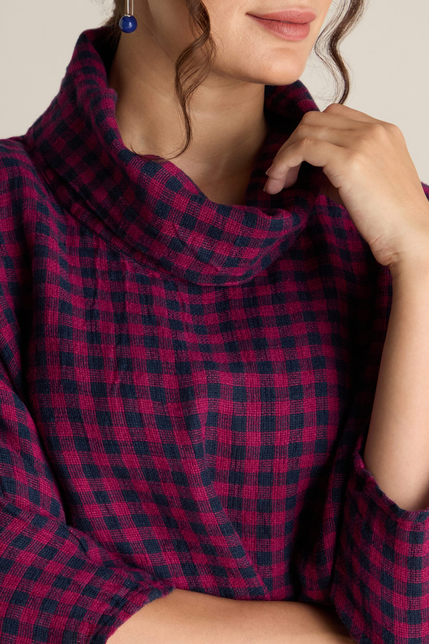 A person wearing the Linen Check Cowl Top, which features a red and blue checkered pattern and a boxy fit, with one hand resting near the neck. They have short, curly hair and are wearing a round blue earring.