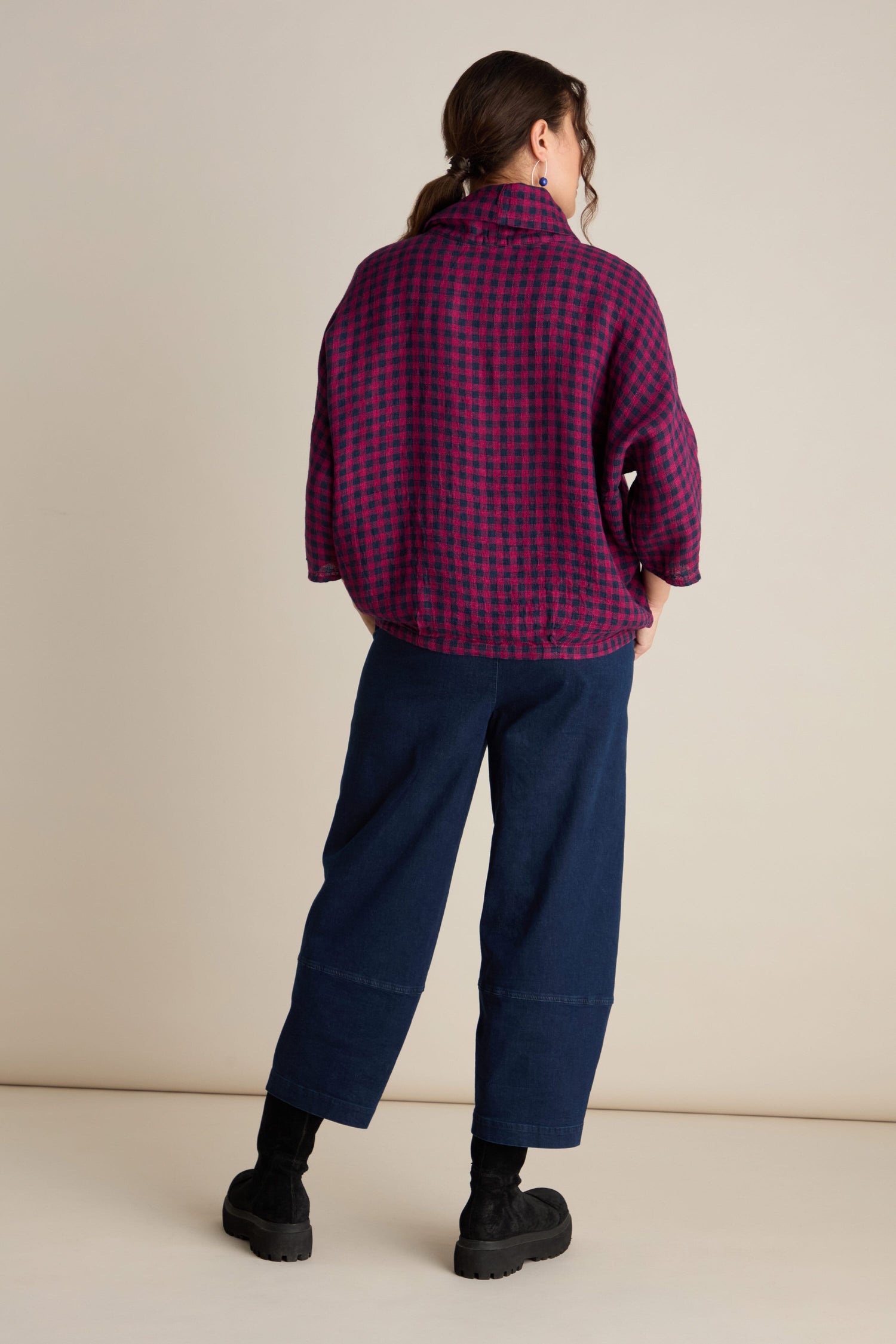 A person stands facing away, sporting a Grizas style with a purple and black Linen Check Cowl Top in a boxy fit, dark blue wide-legged pants, and black boots.