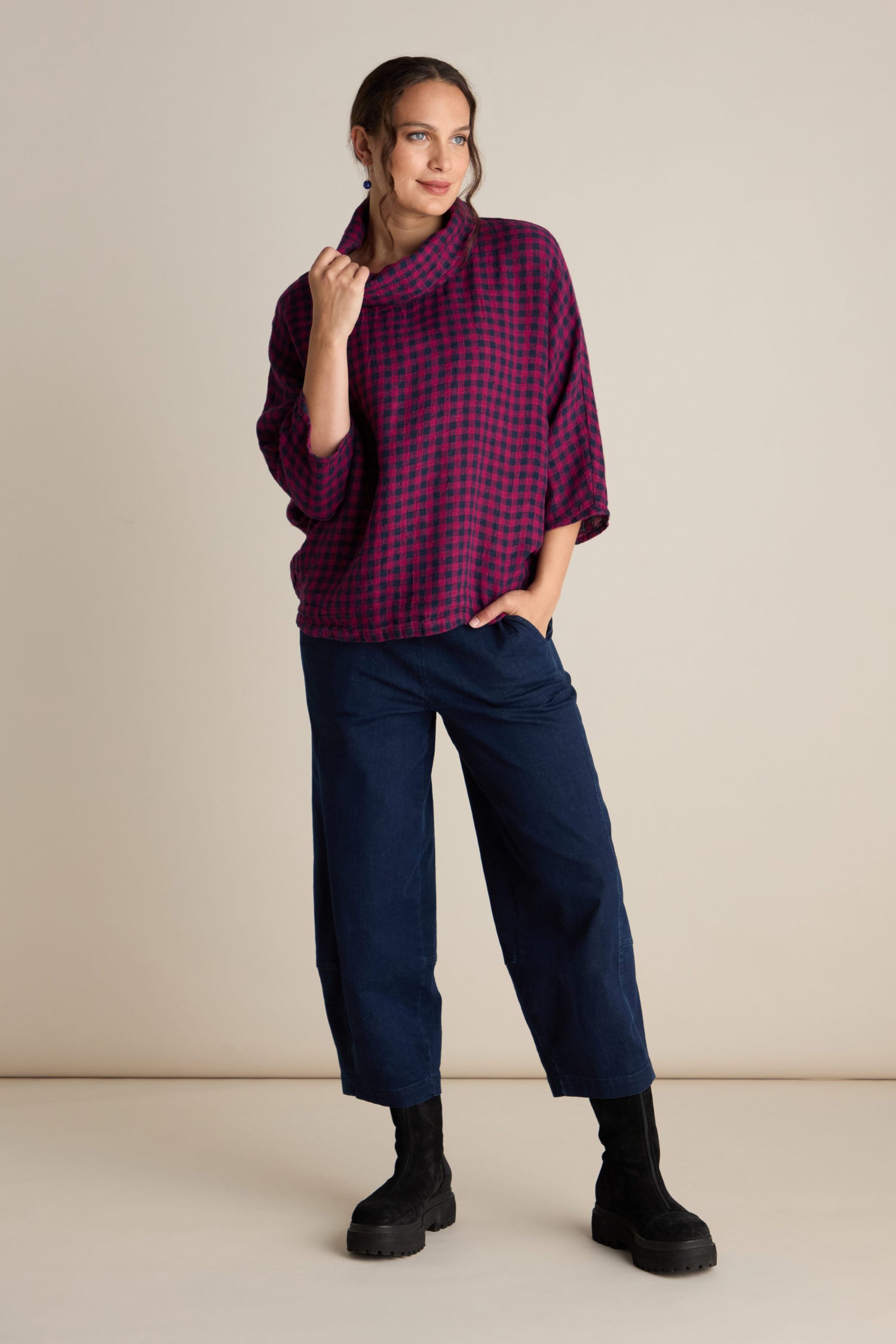 A person stands posing with one hand in their pants pocket, wearing a Linen Check Cowl Top with a boxy fit in purple plaid, dark blue pants, and black boots, against a plain background.