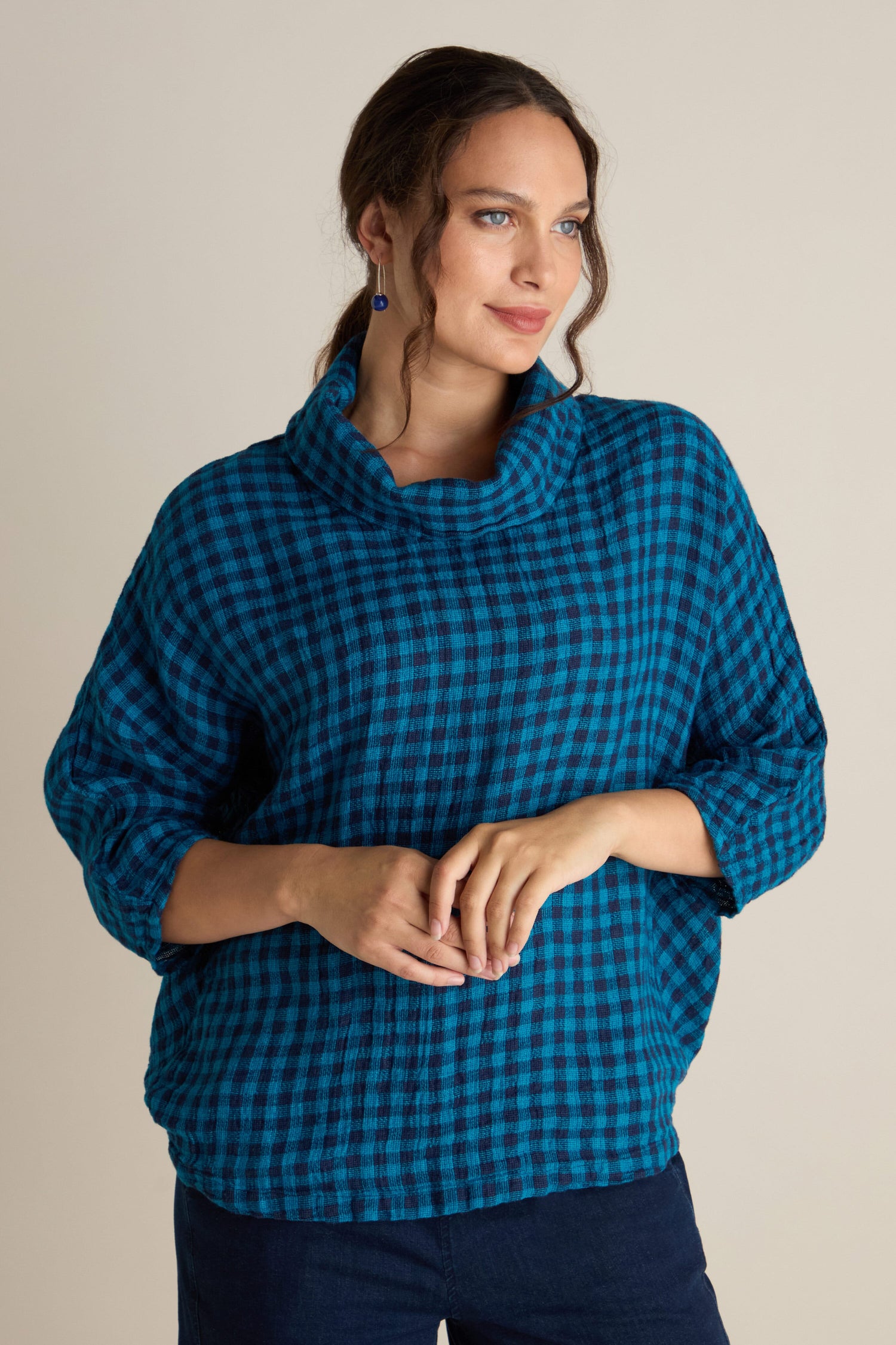 A woman wearing the Linen Check Cowl Top in blue and black posed against a neutral background, gazing to the side. The boxy fit imbues it with a relaxed, Grizas-style essence.