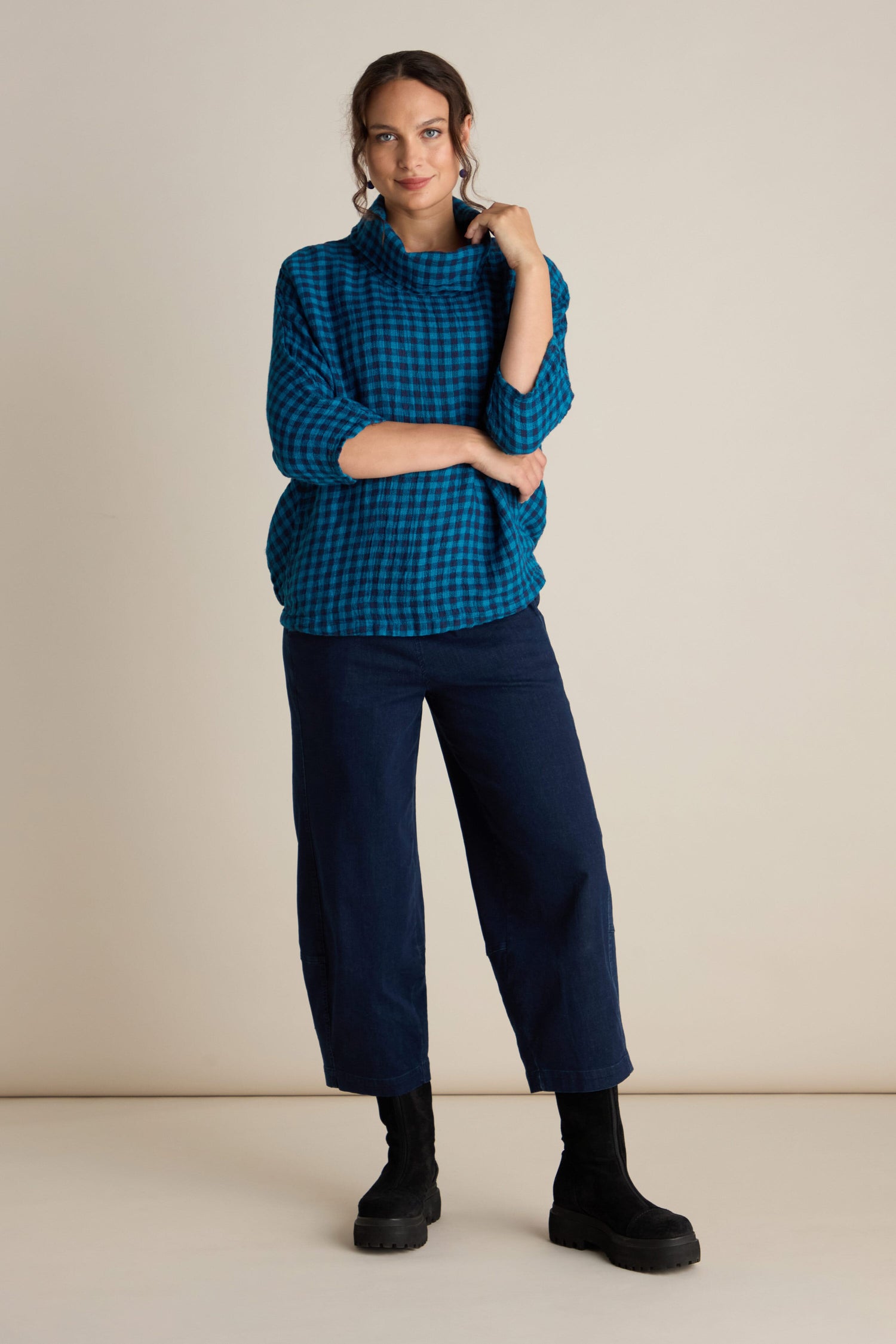 A person stands against a plain background wearing the Linen Check Cowl Top, which features a blue checkered pattern and an oversized fit reminiscent of Grizas style. They pair it with dark pants and black boots. One arm is crossed while the other hand touches the collar of their stylish linen cowl neck top.