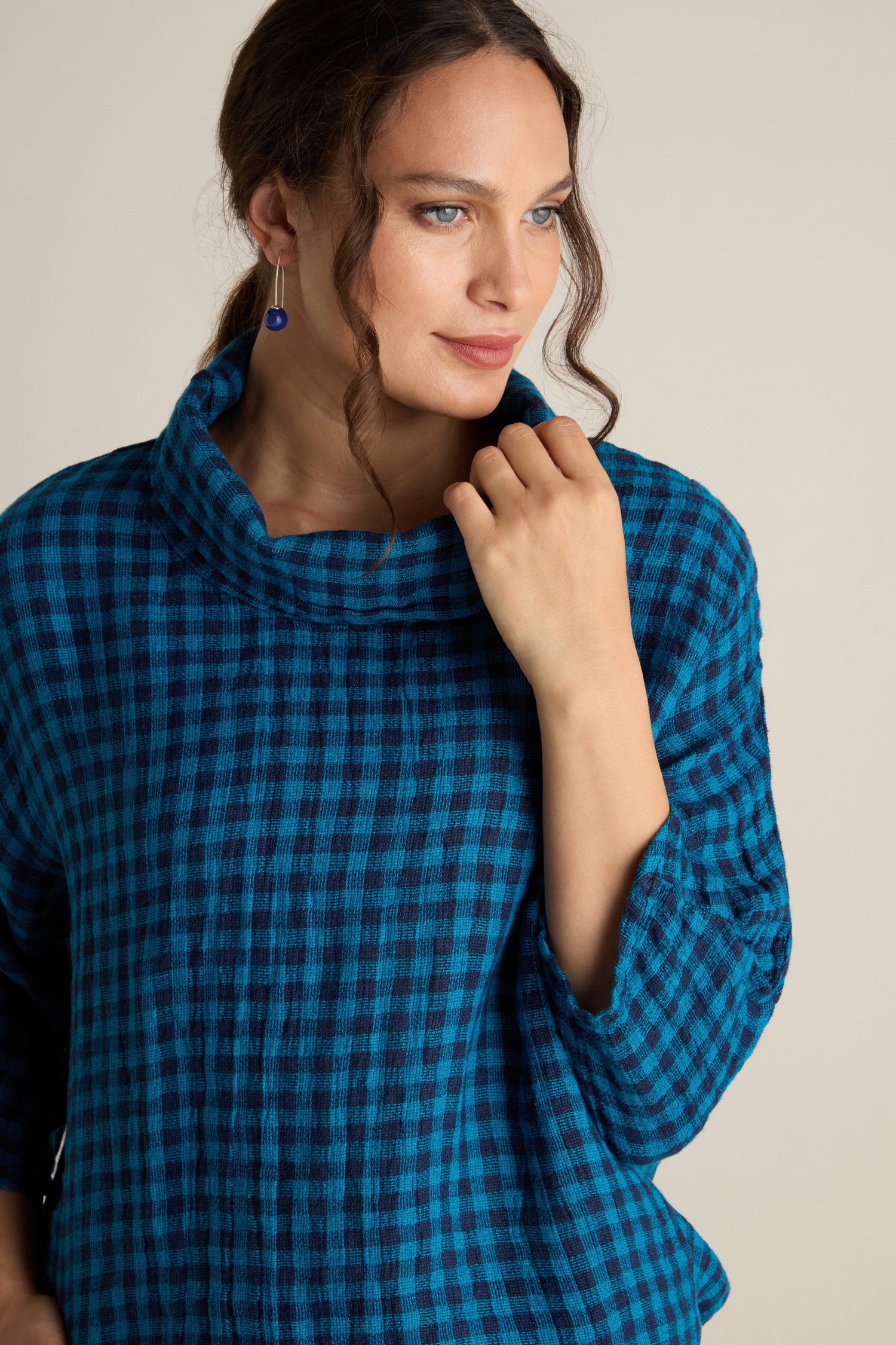 A person with a calm expression, wearing a blue checkered Linen Check Cowl Top and a blue earring, stands against a neutral background.