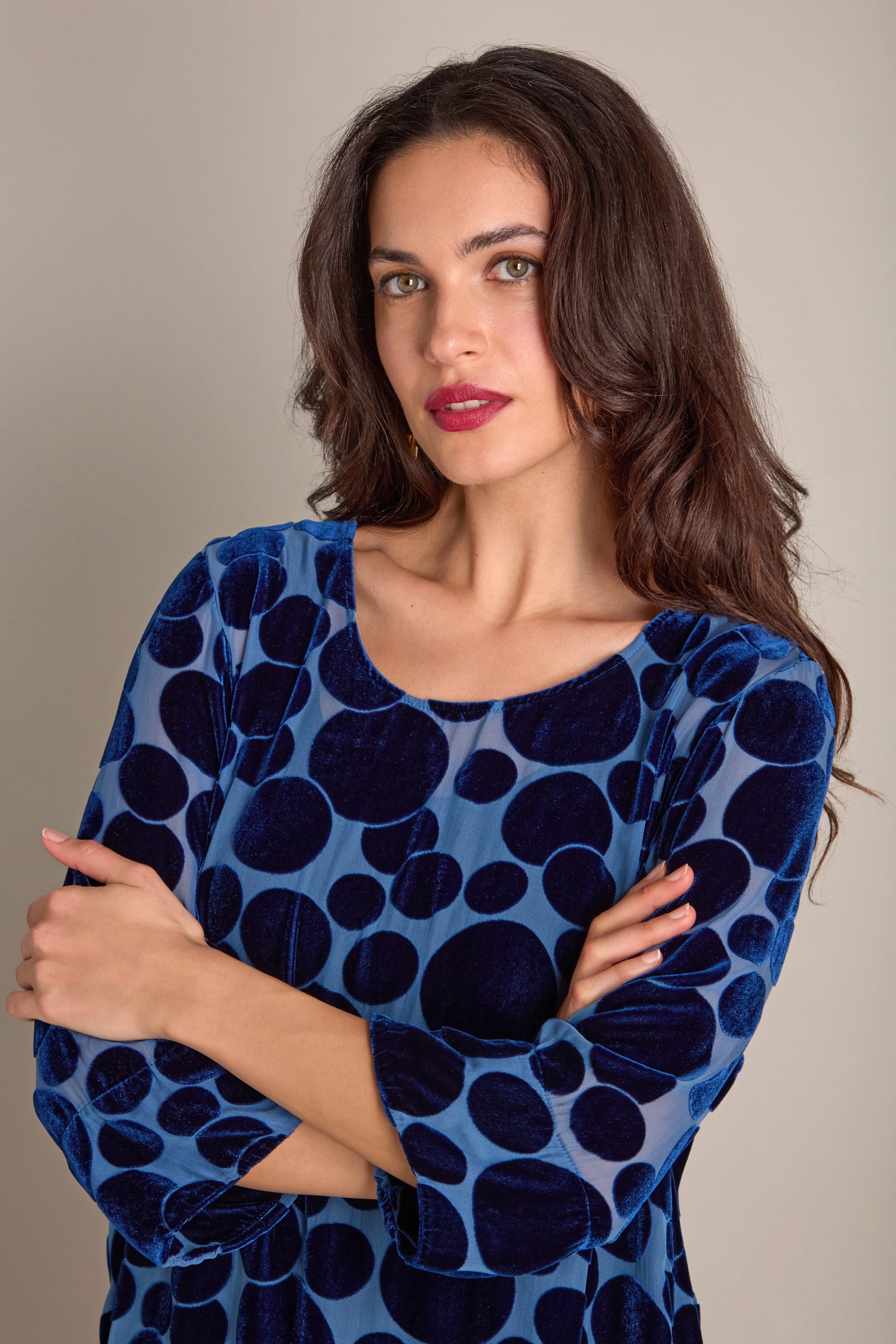 Woman with long brown hair wearing a blue velvet Spot Devore Top with large dark circles, standing against a neutral background with arms crossed.