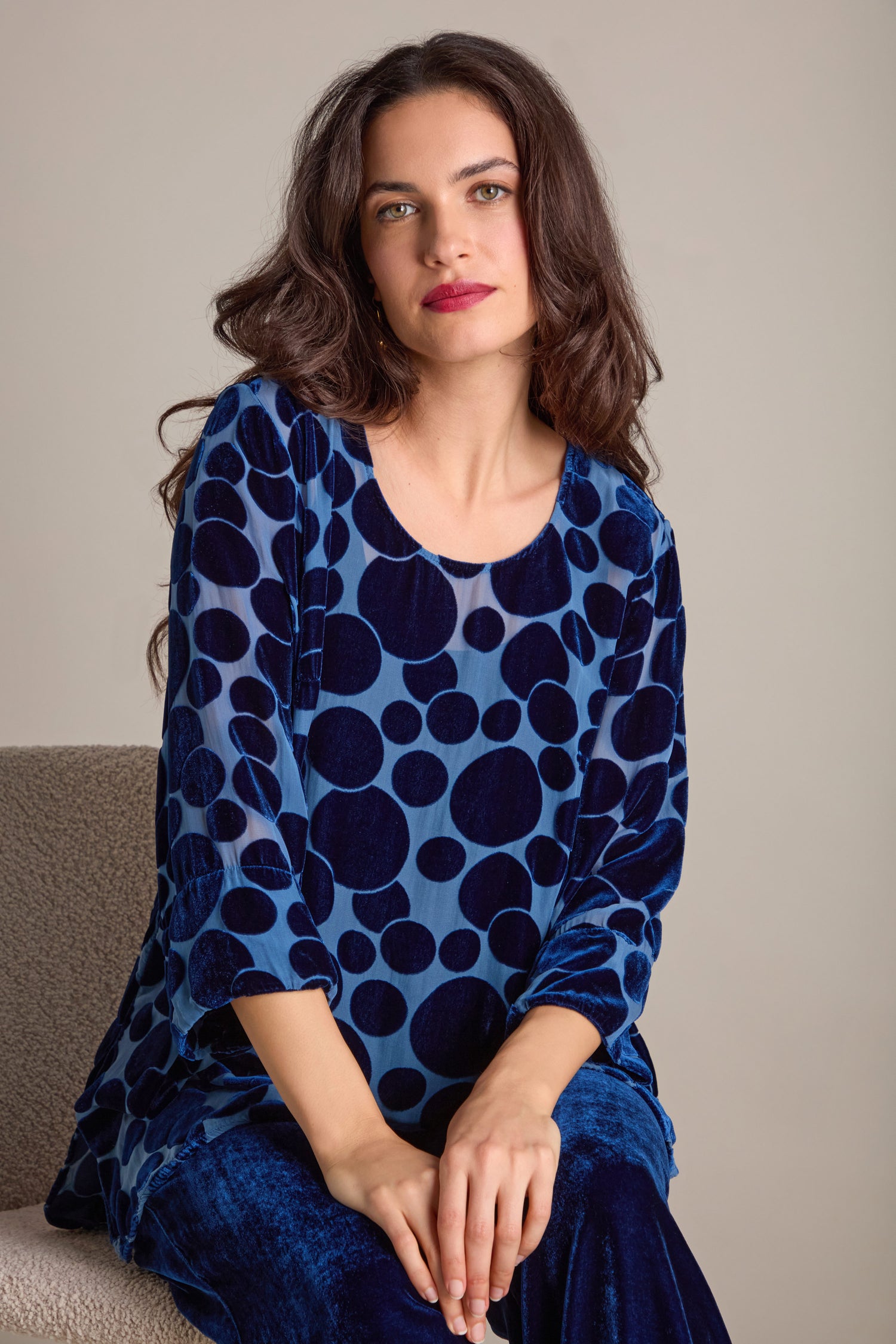 A person with long dark hair sits in a Spot Devore Top featuring sheer detailing, set against a neutral background.