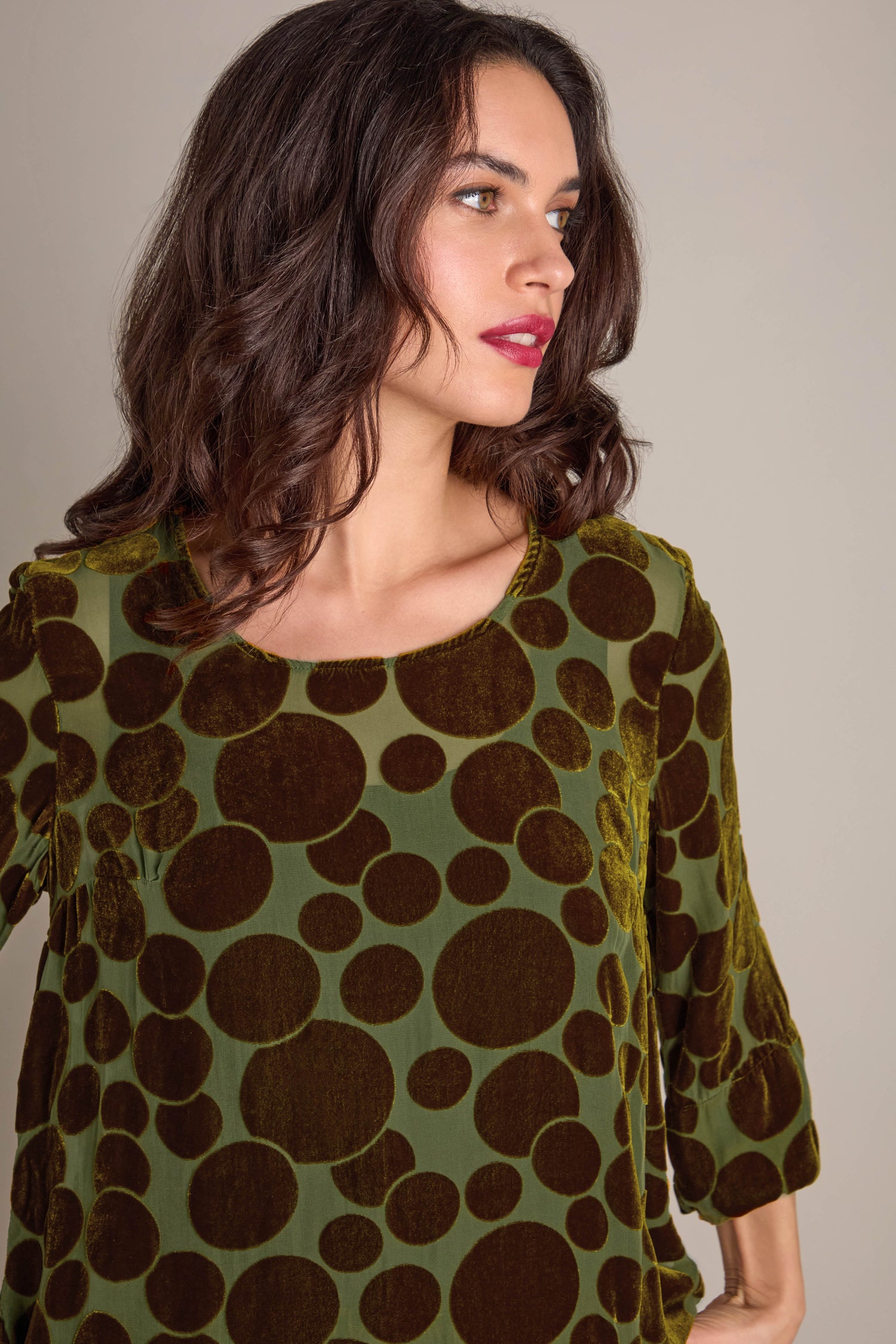 A woman with brown hair is wearing a Spot Devore Top in green velvet, featuring large brown polka dots, as she looks to her left against a plain background.