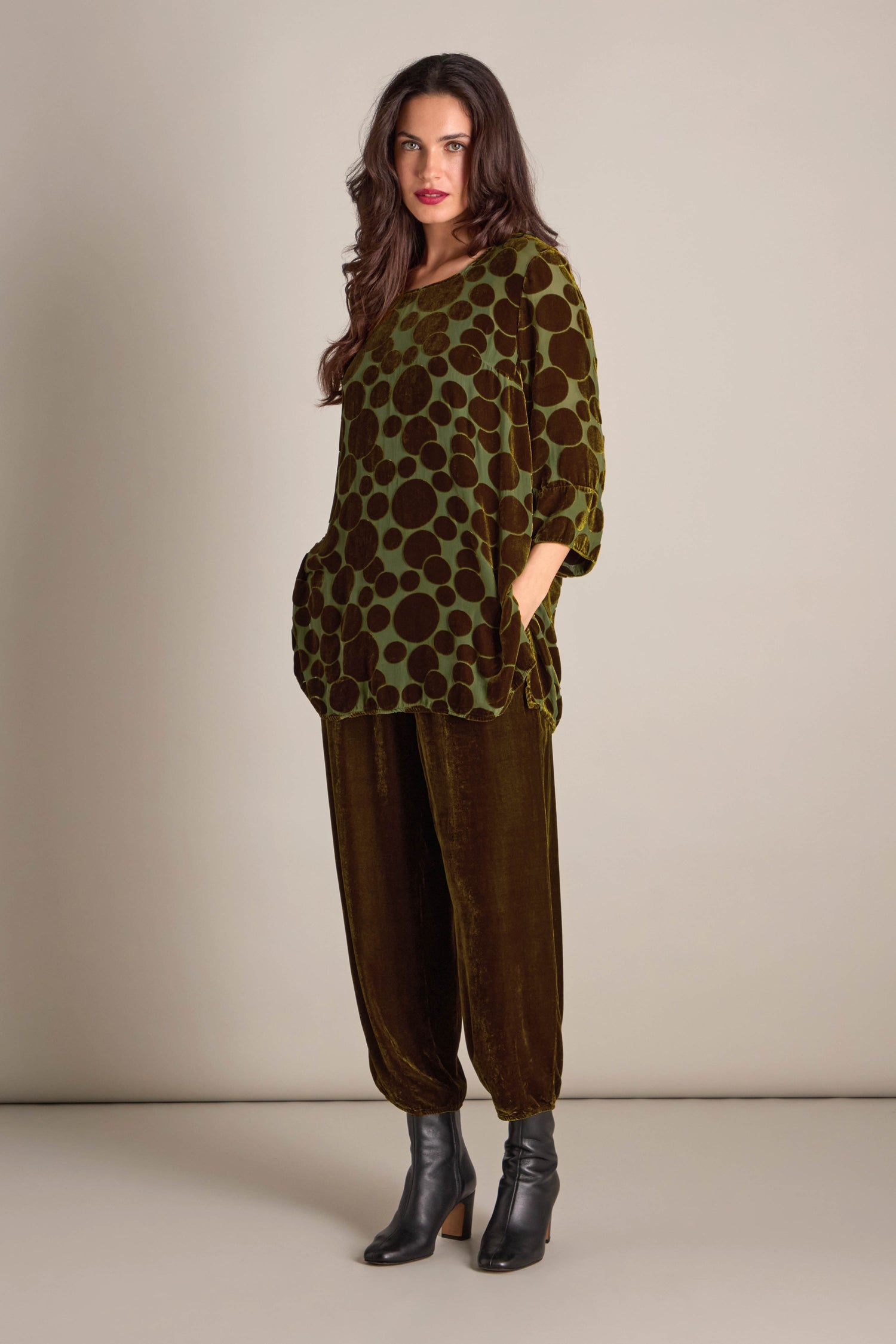 A person stands against a plain background wearing the Spot Devore Top, a green velvet outfit with a polka dot pattern, paired with dark ankle boots.